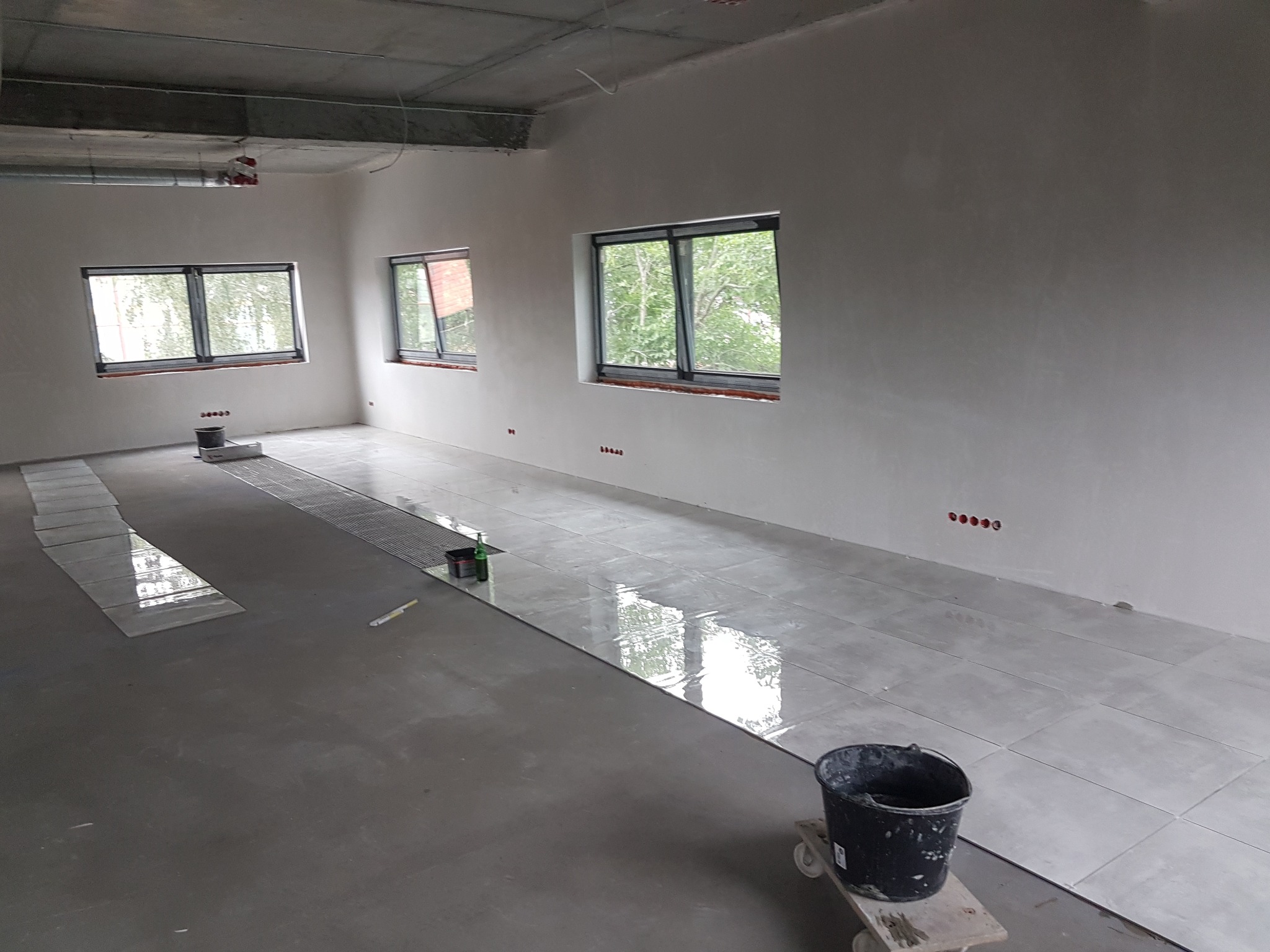 Układanie jasnoszarych płytek podłogowych w dużym, surowym pomieszczeniu z trzema oknami; widoczne wiadro na wózku i narzędzia do cięcia płytek.