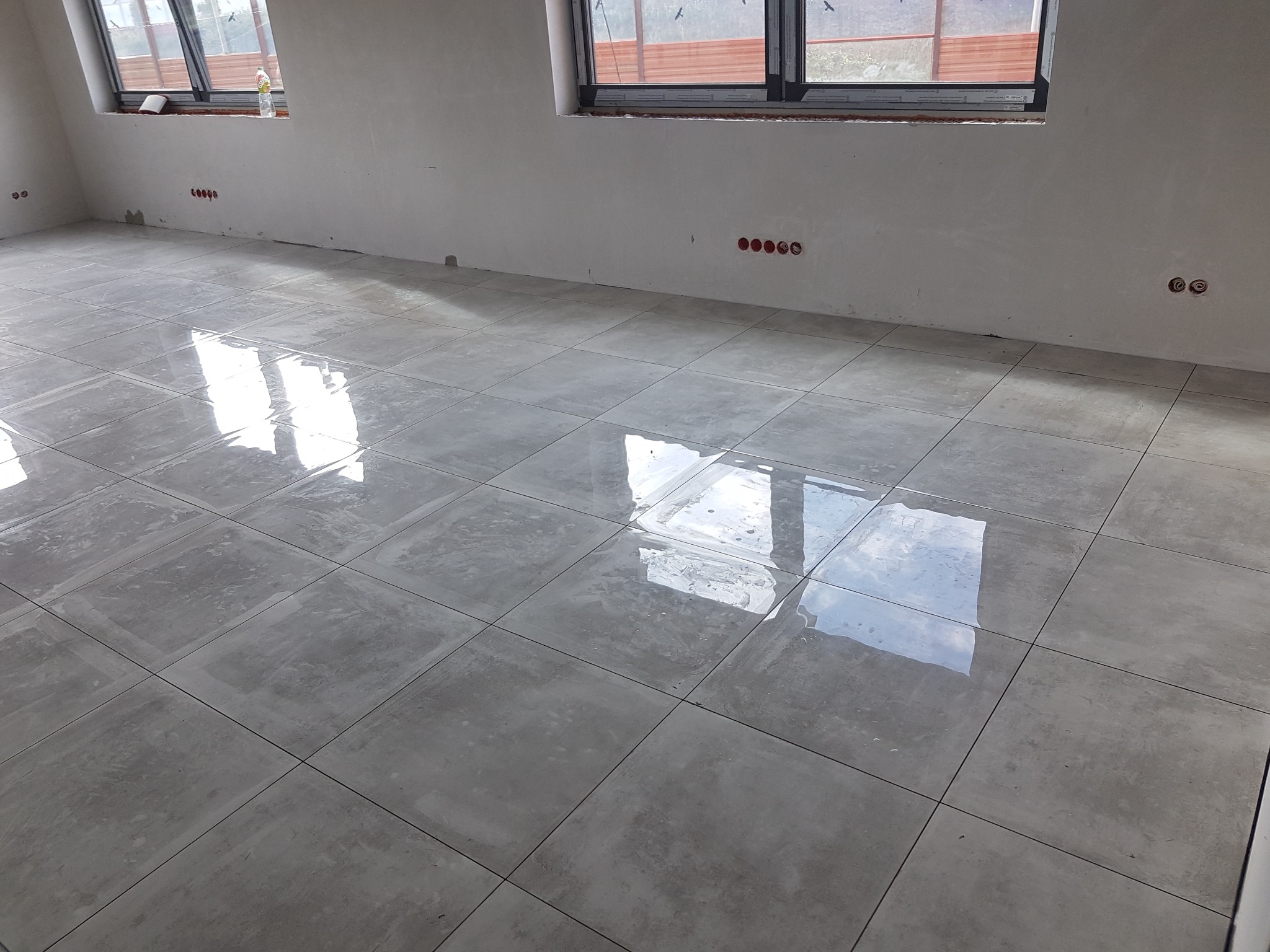 Świeżo ułożone, błyszczące, szare płytki podłogowe w jasnym pomieszczeniu z dwoma oknami i widocznymi puszkami elektrycznymi w ścianie.