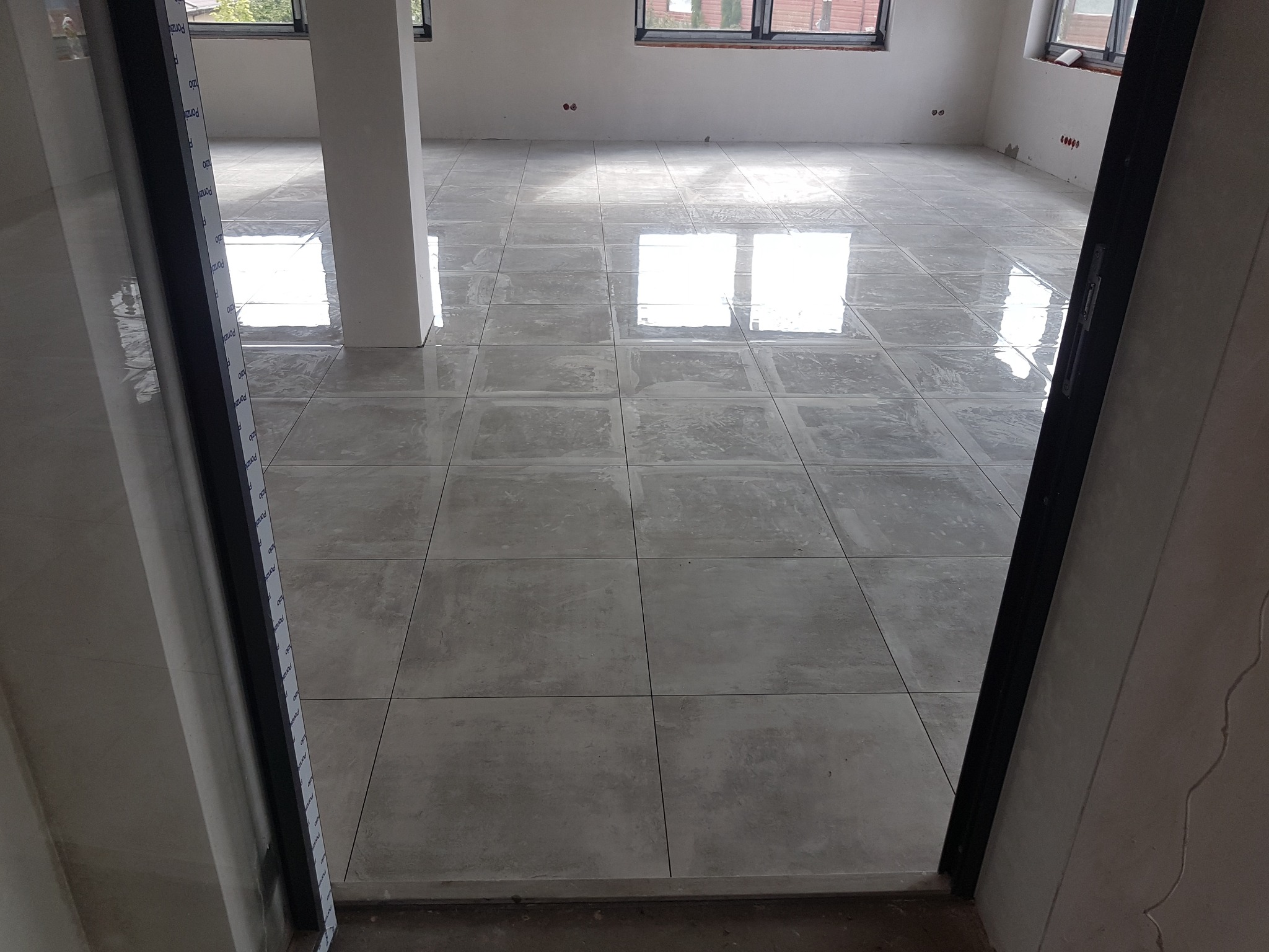 Wykończona podłoga z szarych, kwadratowych płytek w pustym pomieszczeniu, widok zza framugi drzwi. Widoczne okna i białe ściany.