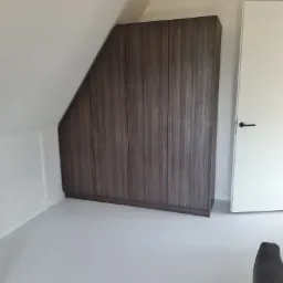 Ciemnobrązowa szafa wnękowa z drewnopodobnym wykończeniem, dopasowana do skośnego sufitu na poddaszu, obok białe drzwi.