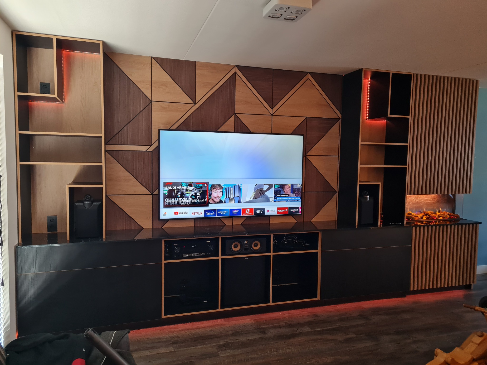 Nowoczesna szafa na wymiar z geometrycznym wzorem na ścianie, podświetleniem LED i miejscem na telewizor oraz sprzęt audio.