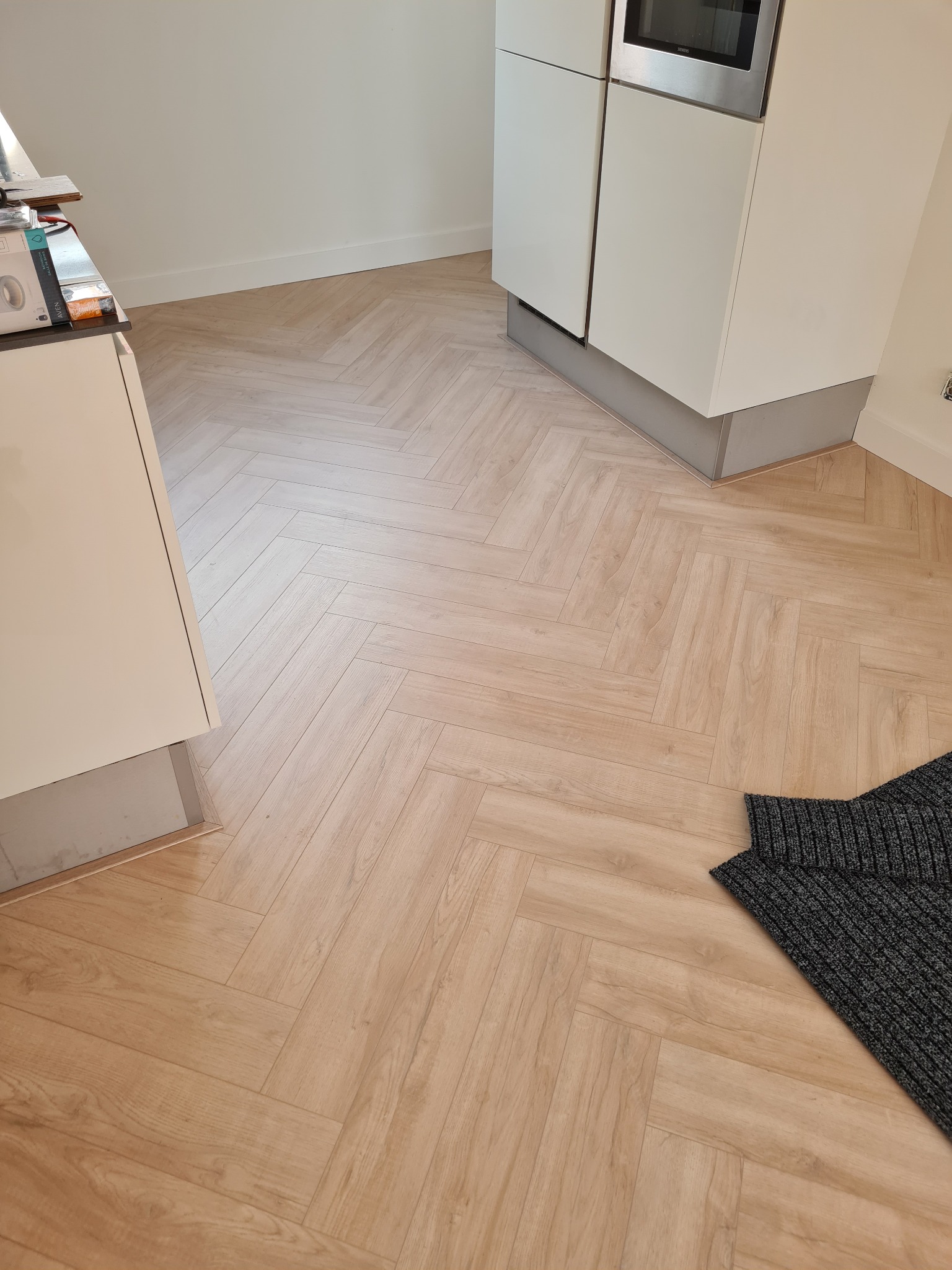 Jasnobrązowe panele ułożone w jodełkę w kuchni z białymi szafkami i szarym dywanikiem w rogu.