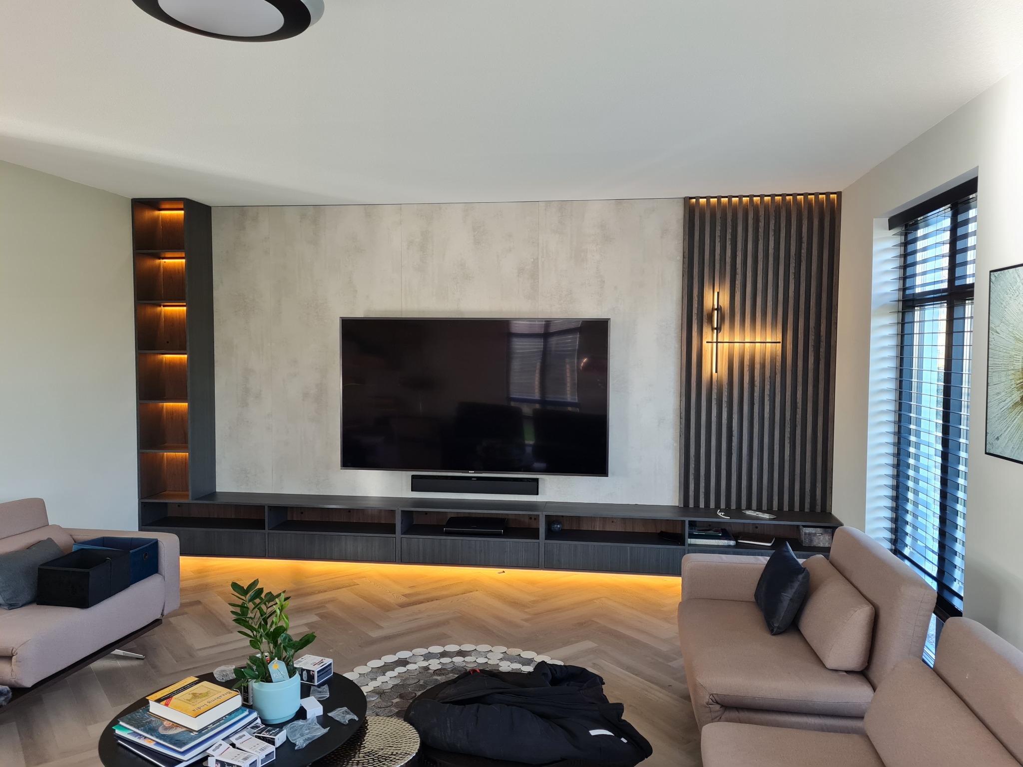 Nowoczesny salon z telewizorem na ścianie, podświetlaną szafką RTV, drewnianymi lamelami dekoracyjnymi z oświetleniem punktowym, oraz regałem z podświetlanymi półkami, uzupełniony o jasne sofy...