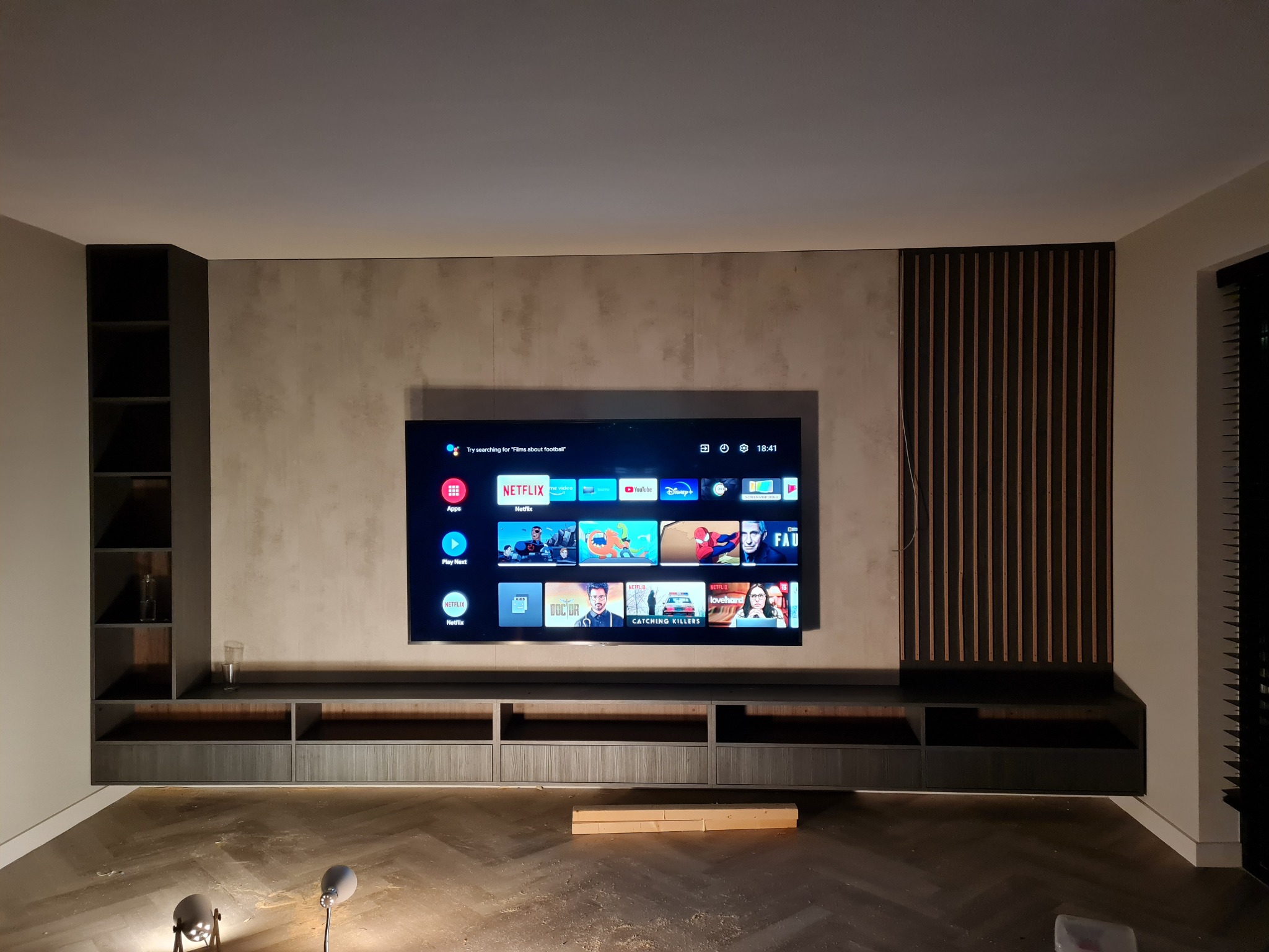 Nowoczesna ściana telewizyjna z podświetlaną szafką RTV w kolorze ciemnego drewna, z półkami po lewej stronie i dekoracyjnymi pionowymi panelami po prawej, z telewizorem wiszącym na ścianie...