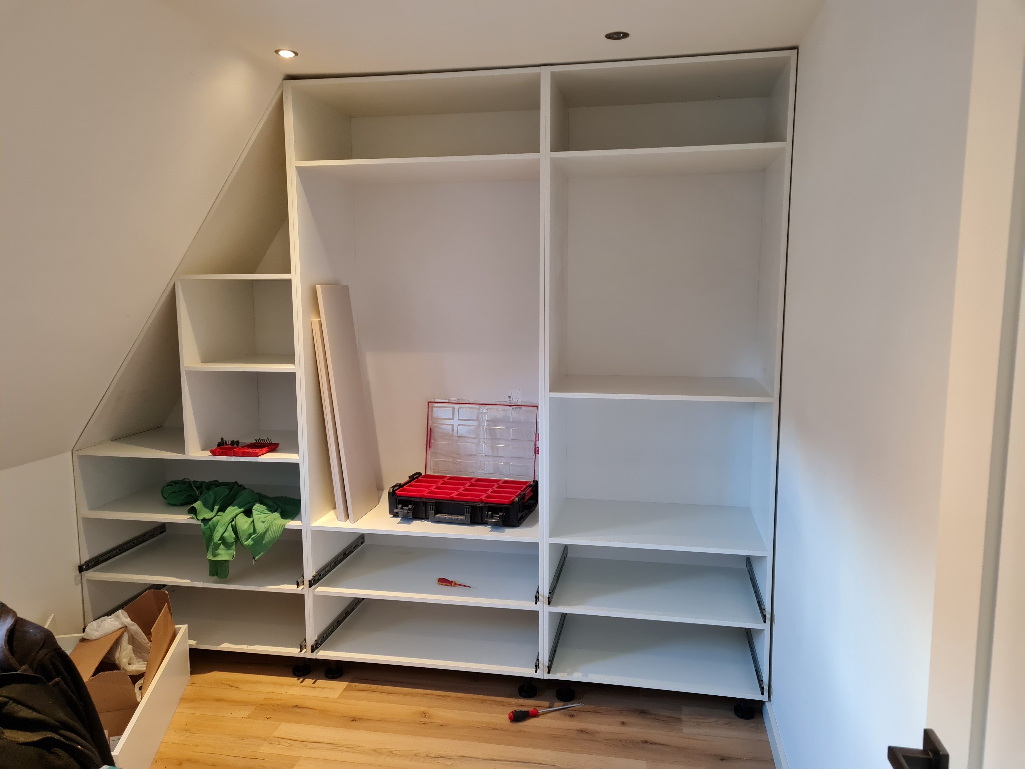 Biała, częściowo zmontowana szafa z szufladami na prowadnicach, widoczna skrzynka z narzędziami, śrubokręt, ubrania i karton na podłodze w trakcie montażu.