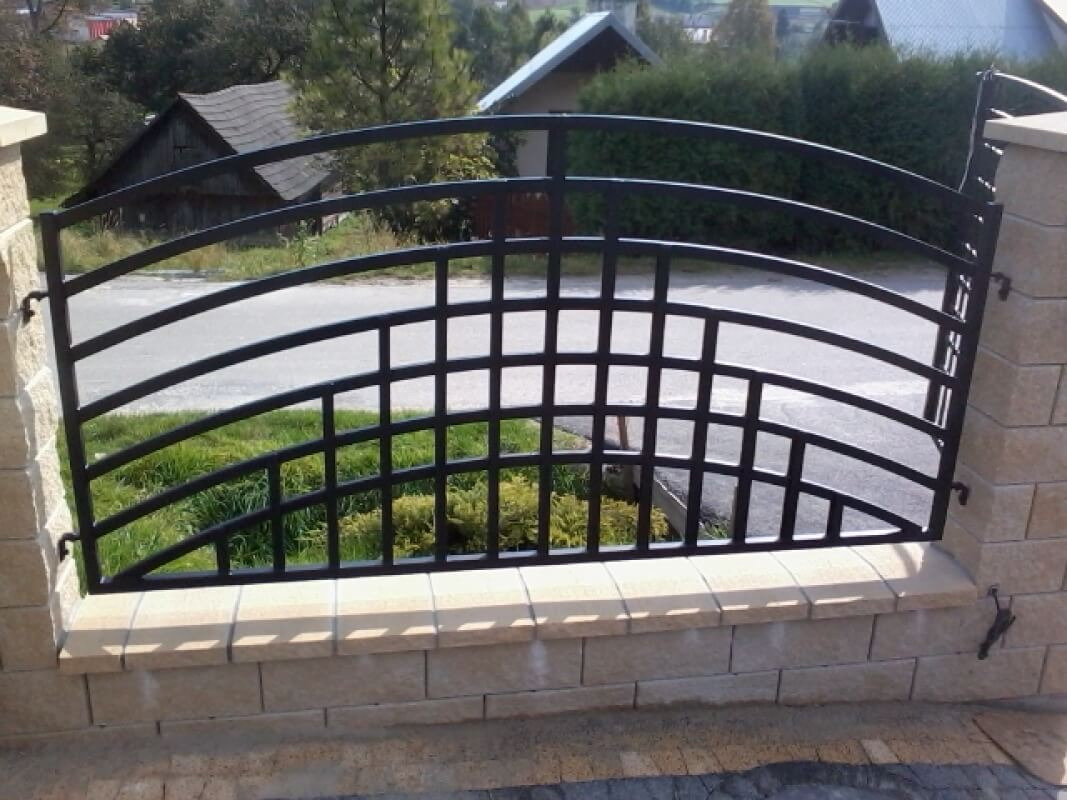 Kute ogrodzenie z dekoracyjnym, łukowatym wypełnieniem, osadzone na niskim murku z kamiennych bloczków, widok na okoliczne domy i zieleń.