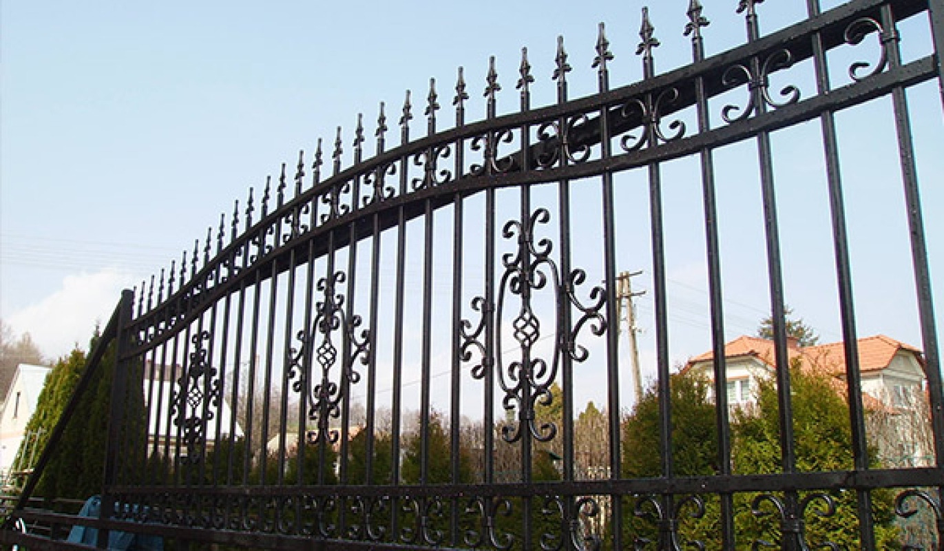 Czarne, ozdobne ogrodzenie z kutego żelaza z widocznymi detalami w kształcie rozet i grotów, na tle domów i zieleni.