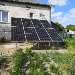 Panele słoneczne zamontowane na metalowych wspornikach w przydomowym ogr&oacute;dku warzywnym, na tle białego, ceglanego domu i błękitnego nieba z chmurami.