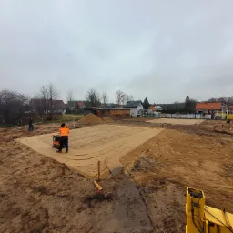Pracownik w pomarańczowej kamizelce ubija piasek zagęszczarką na placu budowy, widoczne przygotowane obszary pod fundamenty w kształcie prostokątów, w tle zabudowa jednorodzinna i pochmurne niebo.