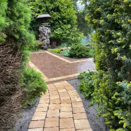 Kremowa kostka brukowa ułożona w krętą ścieżkę ogrodową, otoczona zielenią krzewów i drzew, z kamienną studnią po lewej stronie.