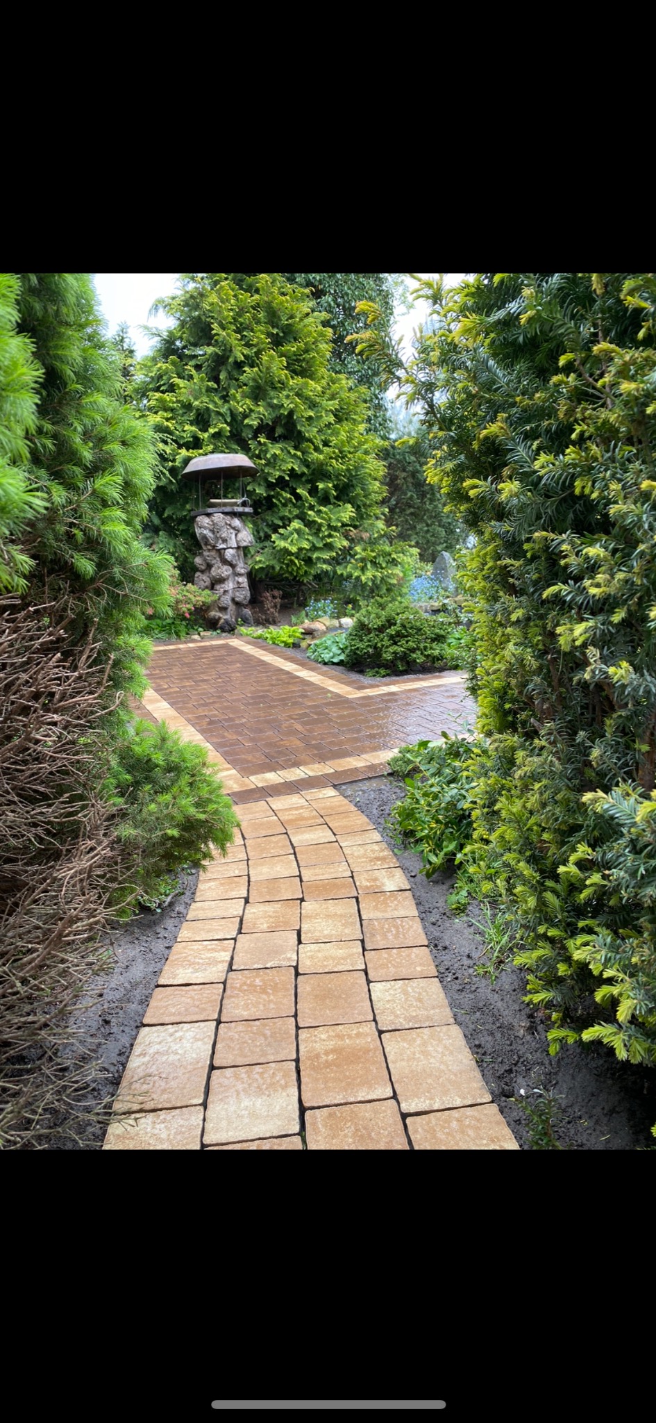 Kremowa kostka brukowa ułożona w krętą ścieżkę ogrodową, otoczona zielenią krzewów i drzew, z kamienną studnią po lewej stronie.