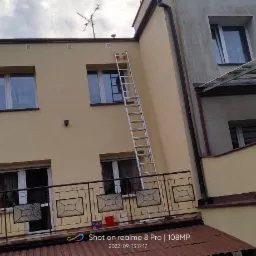 Żółta elewacja budynku z aluminiową drabiną opartą o ścianę, widok z dołu, balkon z metalową balustradą na pierwszym planie.