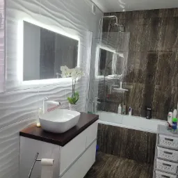 Nowoczesna łazienka z białymi, falistymi płytkami na ścianie, podświetlanym lustrem, umywalką nablatową i brązowymi płytkami imitującymi marmur na podłodze oraz ścianie prysznica.