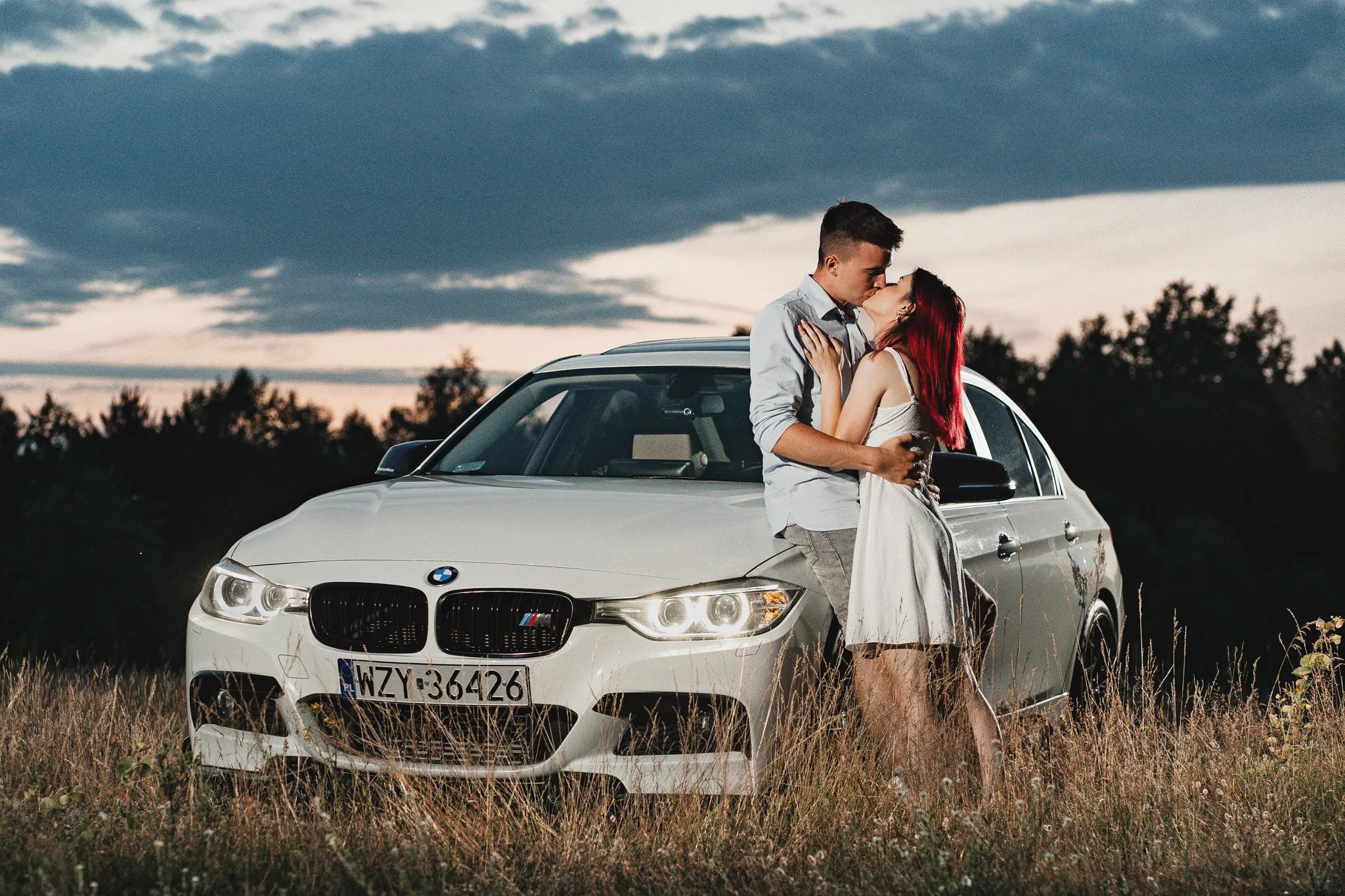 Zakochana para całuje się na tle białego BMW z widoczną tablicą rejestracyjną, w otoczeniu wysokiej trawy i zachodzącego słońca.