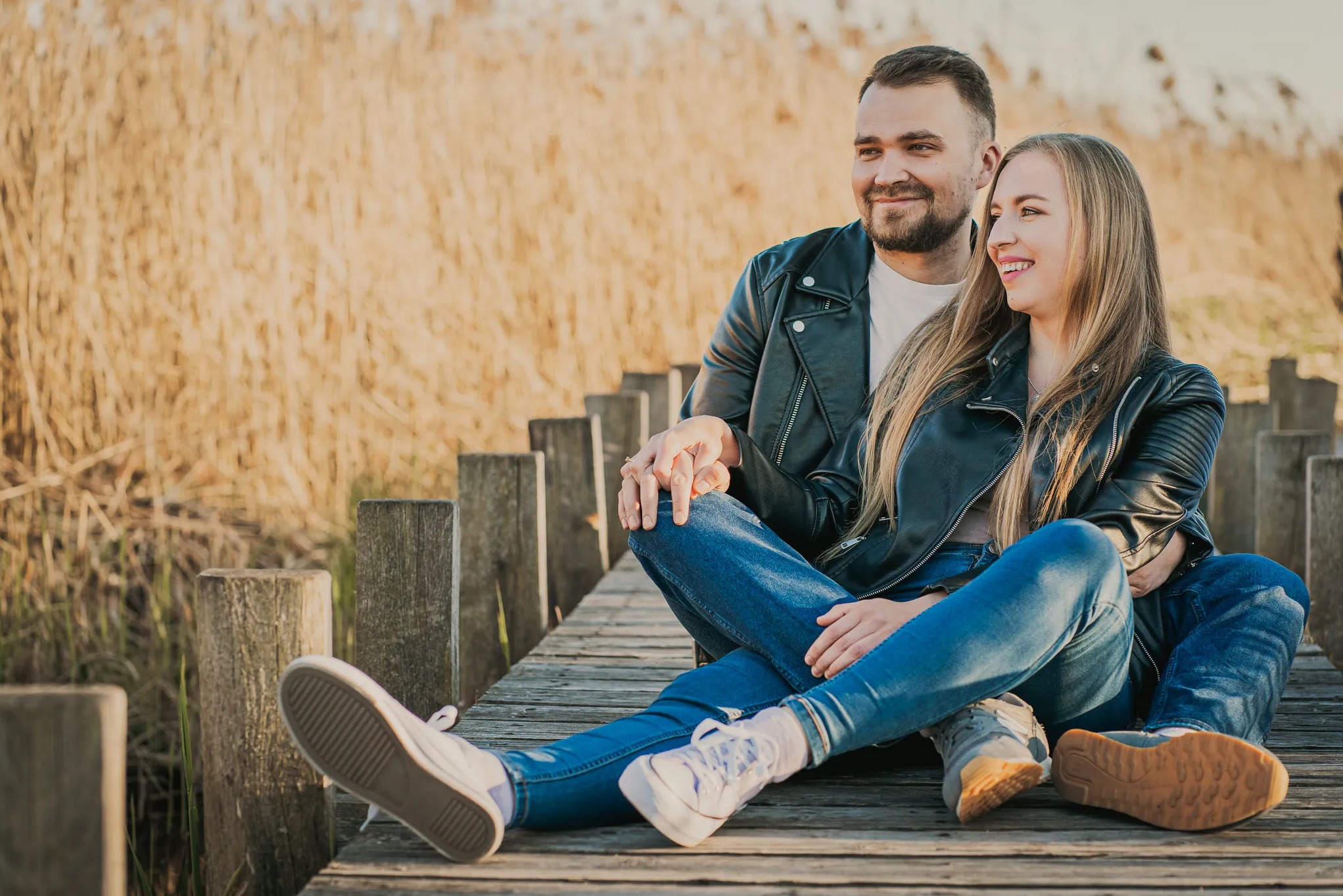 Para w skórzanych kurtkach siedzi na drewnianym pomoście, trzymając się za ręce, z wysoką trawą w tle, w ciepłym, słonecznym świetle.