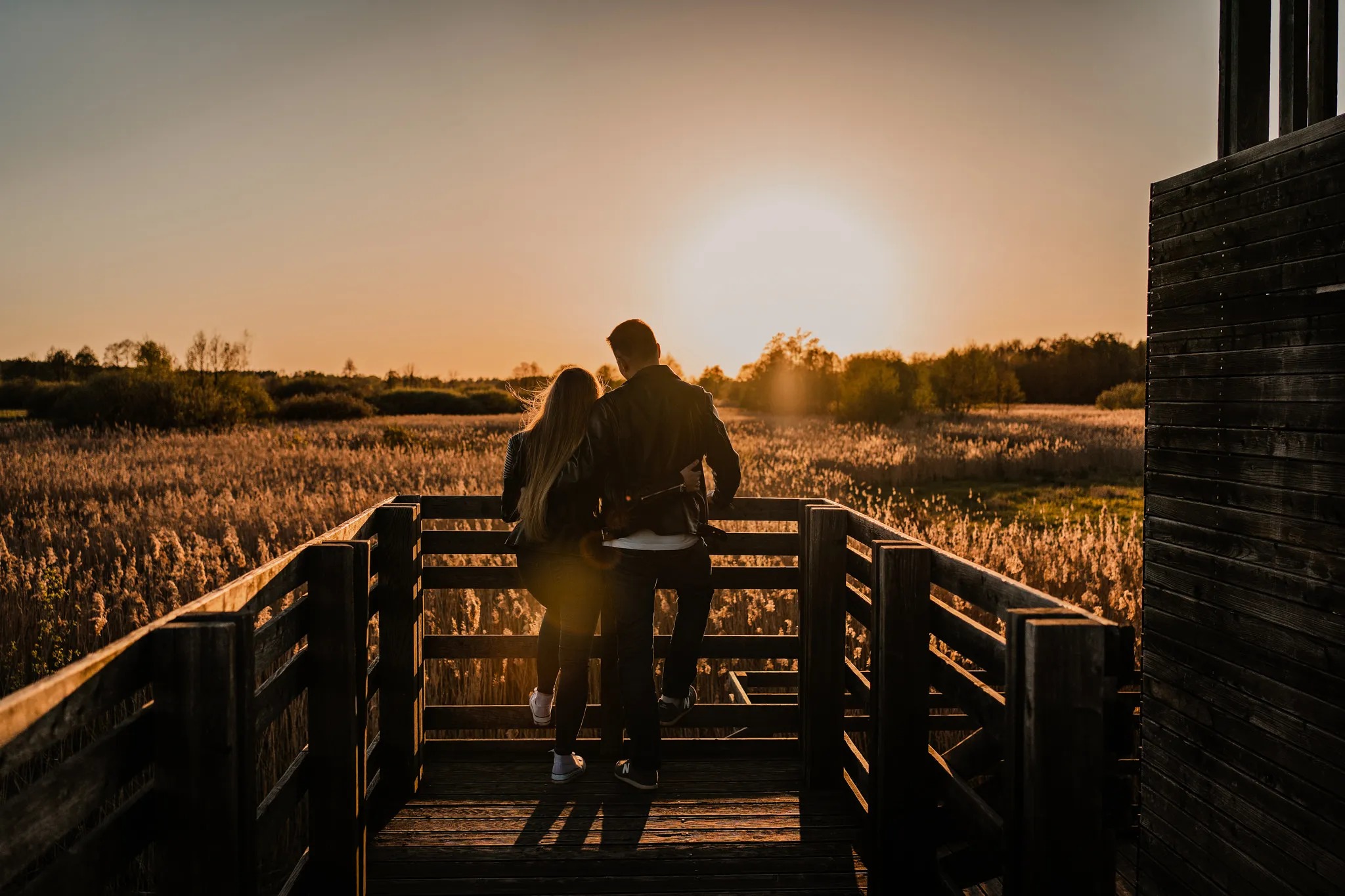 Para, tyłem do widza, obejmuje się na drewnianym pomoście widokowym na tle zachodzącego słońca i łąki trzcinowej.