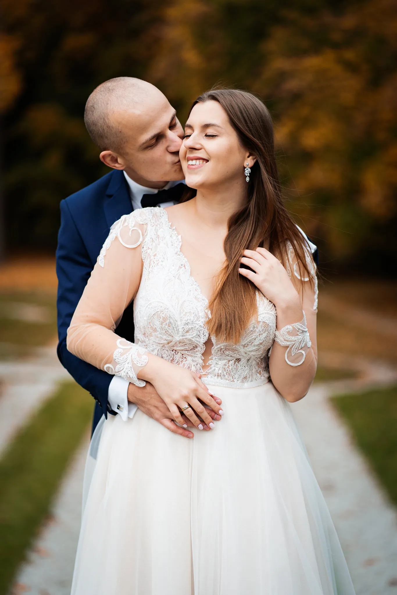 Uśmiechnięta panna młoda w koronkowej sukni z długim rękawem, obejmowana przez pana młodego w granatowym garniturze, który całuje ją w policzek na tle jesiennego parku.