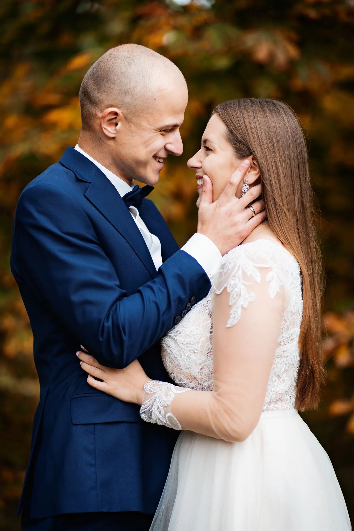 Uśmiechnięta para młoda w plenerze: on w granatowym garniturze z muchą, ona w białej sukni z koronkowymi rękawami i błyszczącymi kolczykami, delikatnie dotykana przez niego po twarzy.