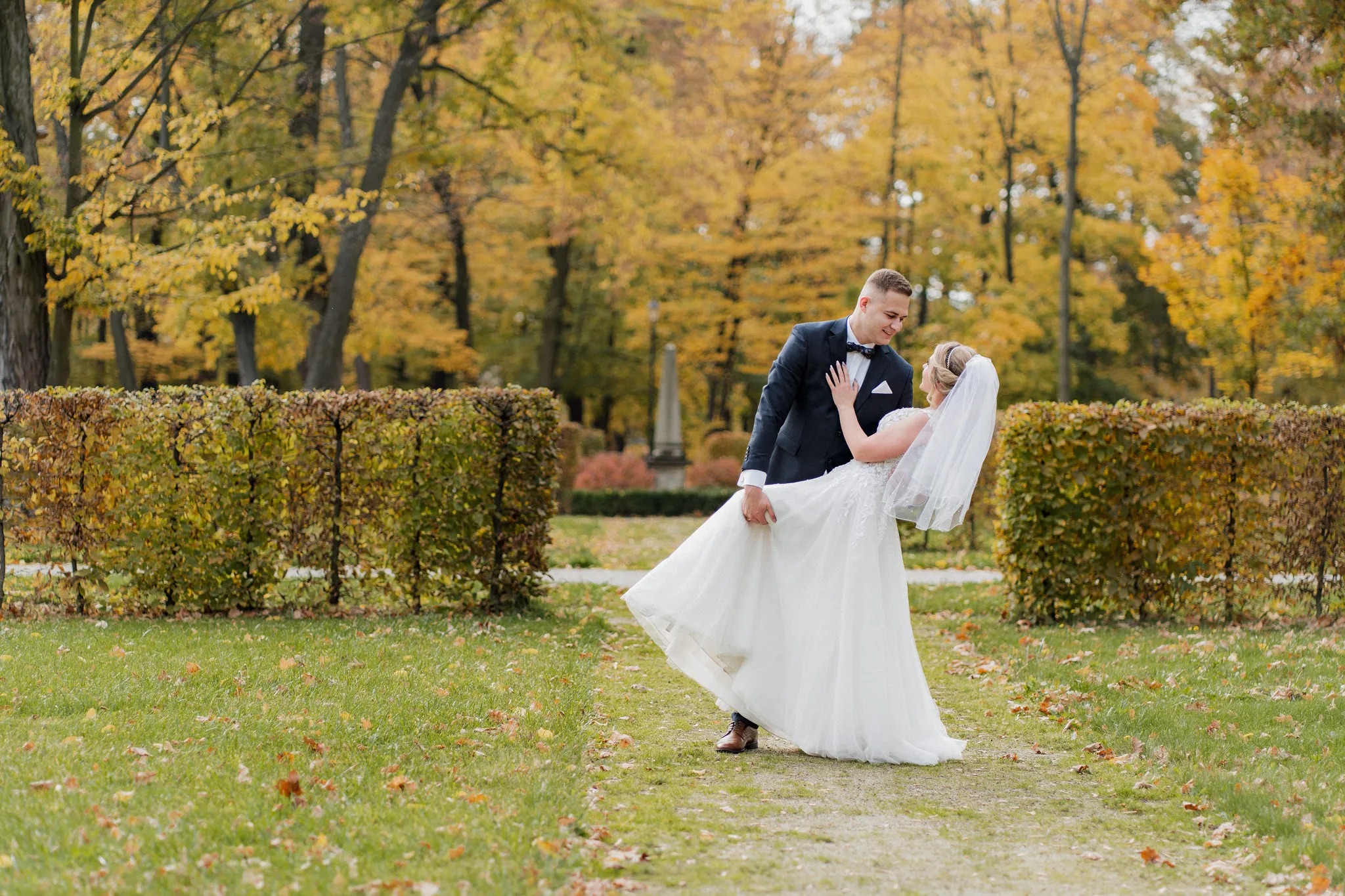Para młoda w jesiennej scenerii parku: pan młody w granatowym smokingu trzyma pannę młodą w białej sukni ślubnej z welonem.