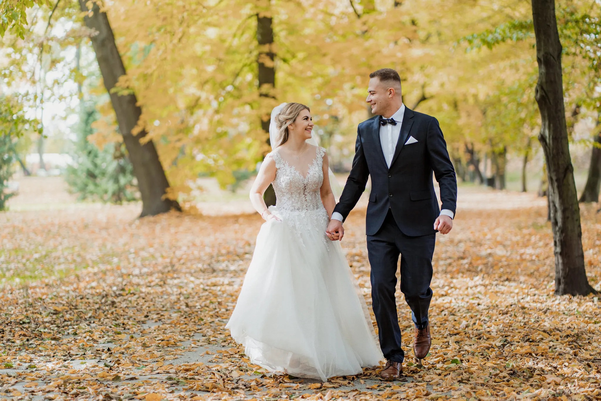 Uśmiechnięta para w strojach ślubnych spaceruje aleją jesiennych drzew, trzymając się za ręce.