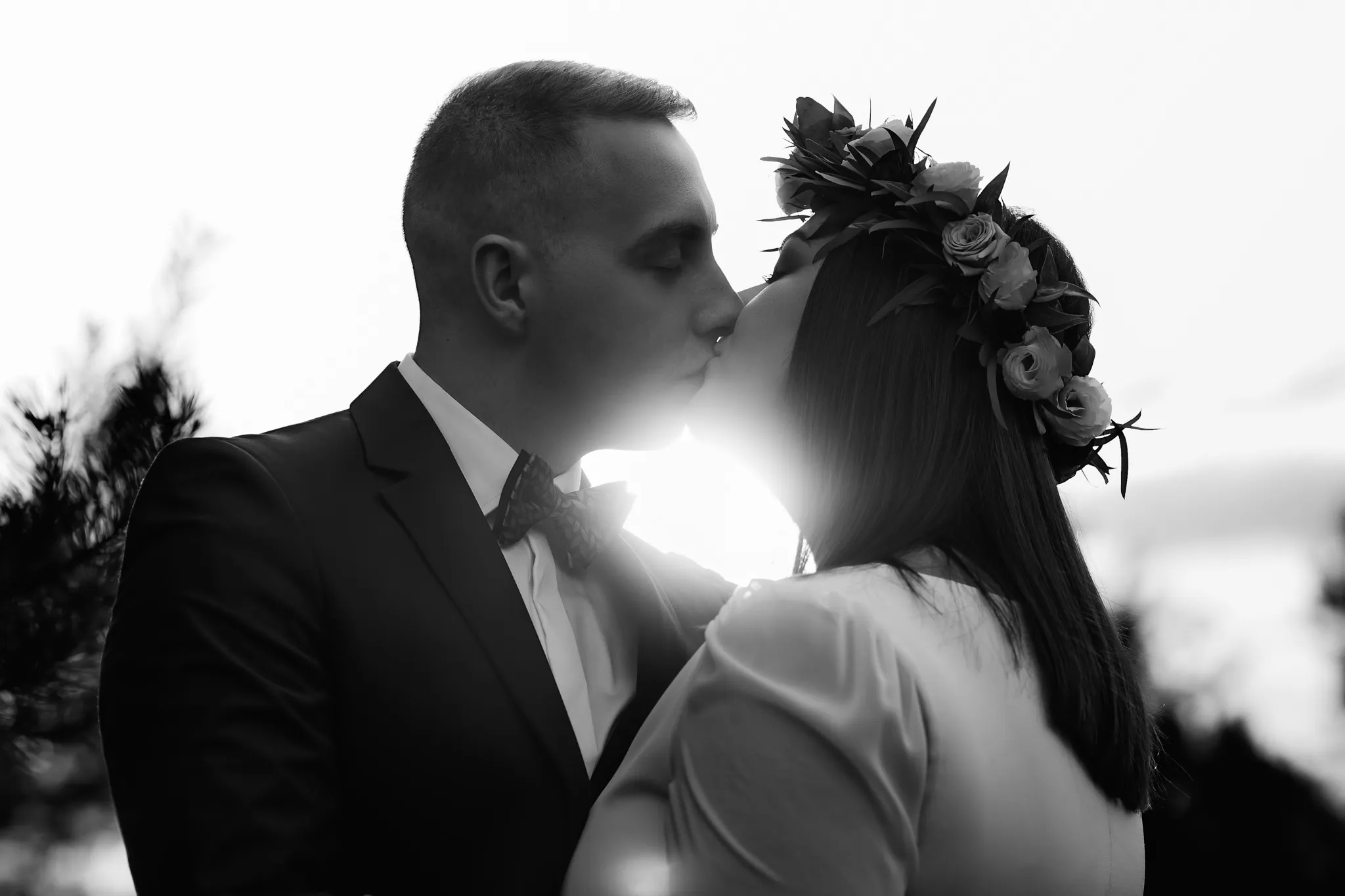 Czarno-białe zdjęcie całującej się pary młodej, panna młoda ma wianek z róż na głowie, a pan młody ma ciemny garnitur i wzorzystą muszkę.