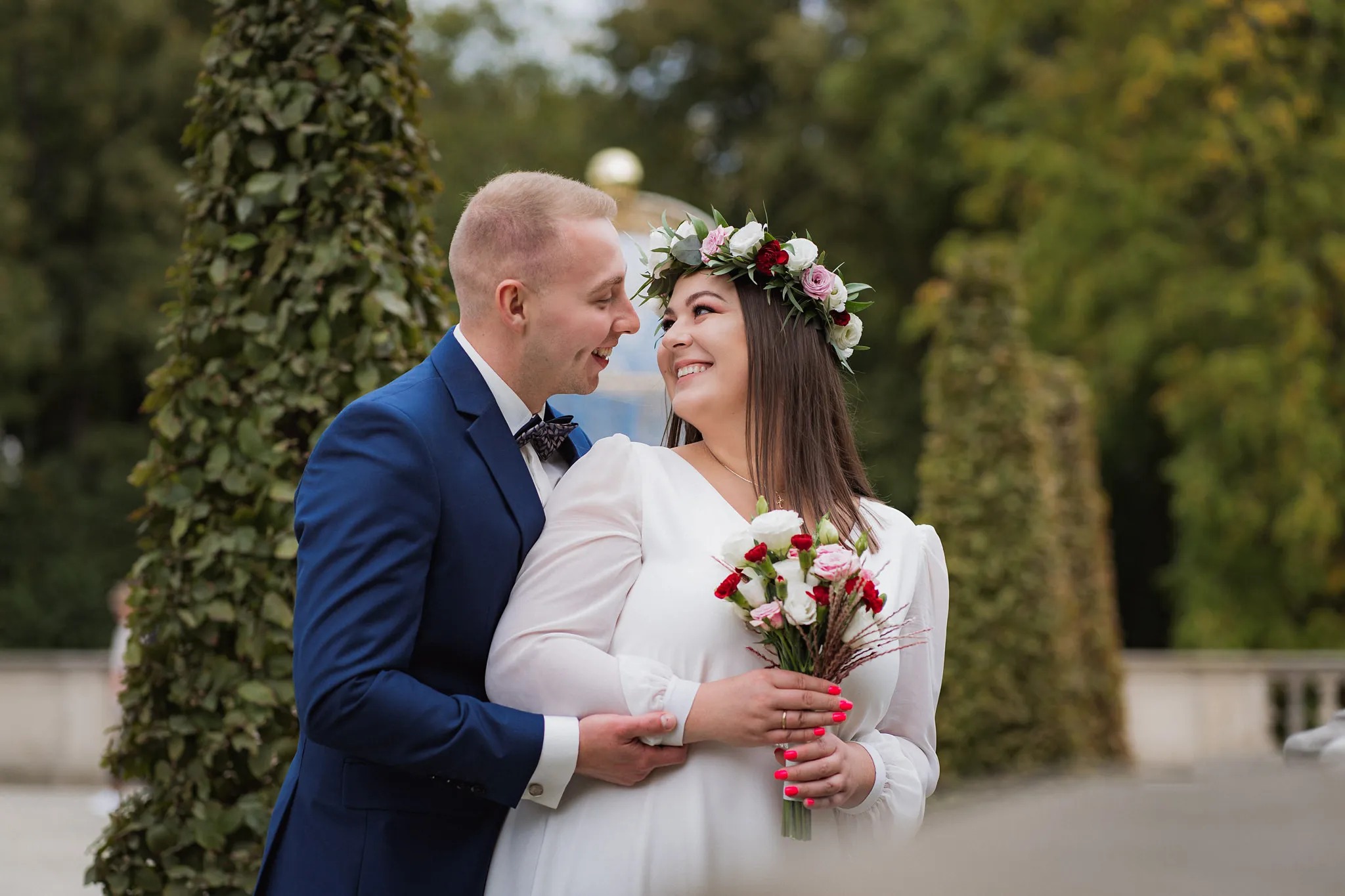 Uśmiechnięta para, panna młoda z wiankiem i bukietem, pan młody w granatowym garniturze, obejmująca się na tle zieleni.