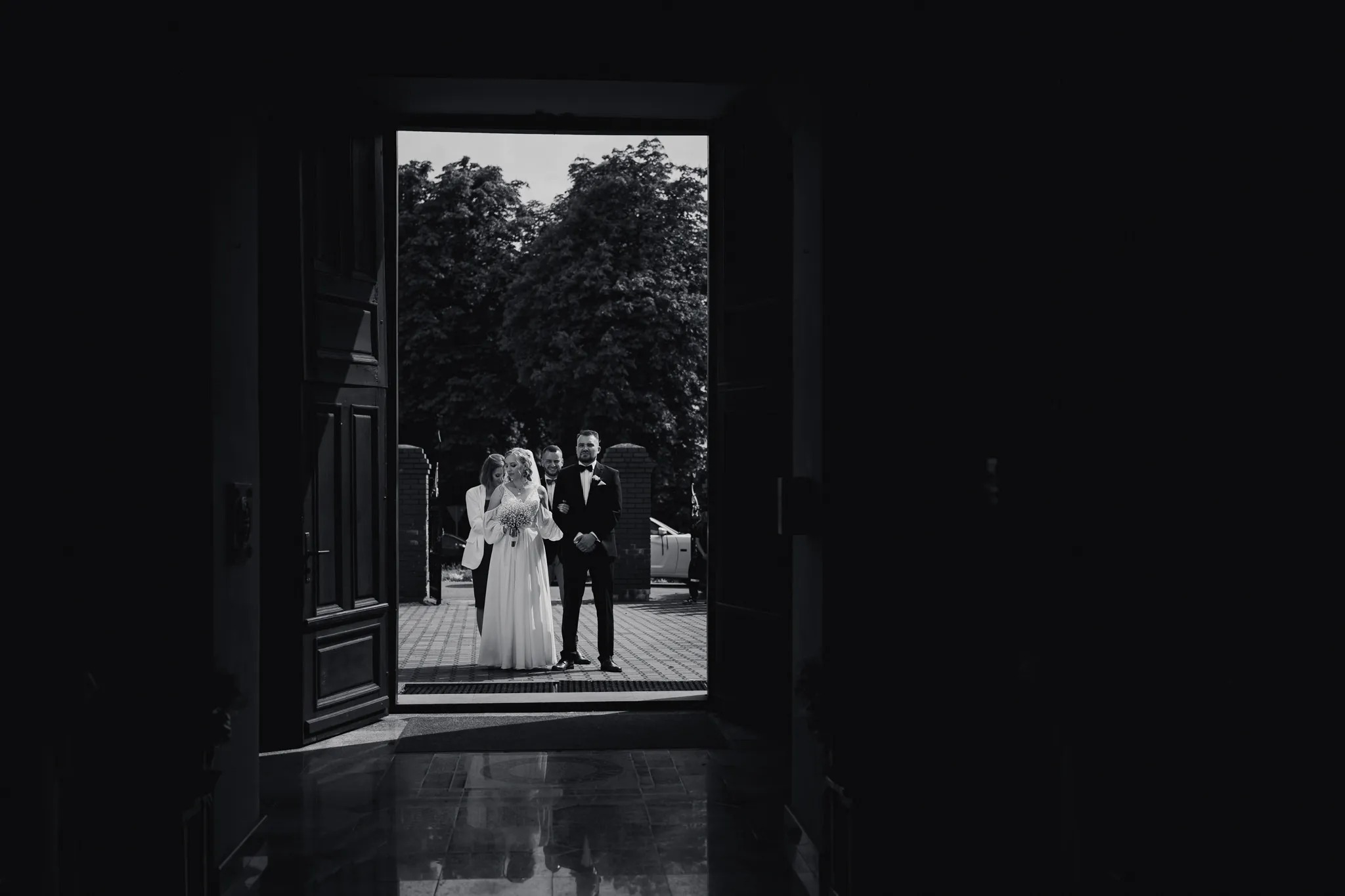 Czarno-białe zdjęcie pary młodej wychodzącej z kościoła, prowadzona przez mężczyznę w smokingu, z gośćmi za nimi, widoczne z ciemnego wnętrza