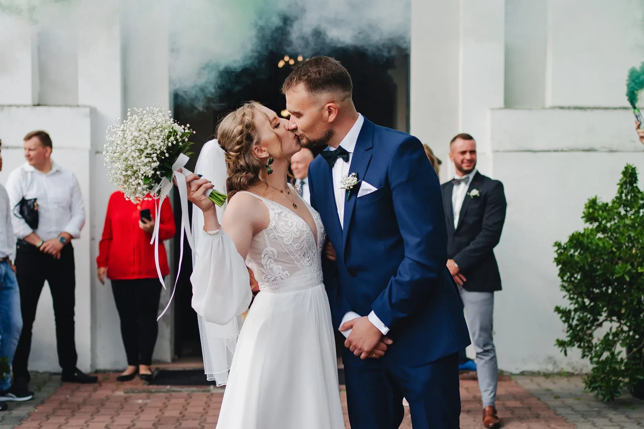 Pocałunek nowożeńców przed kościołem, otoczonych gośćmi i zielonym dymem z rac.
