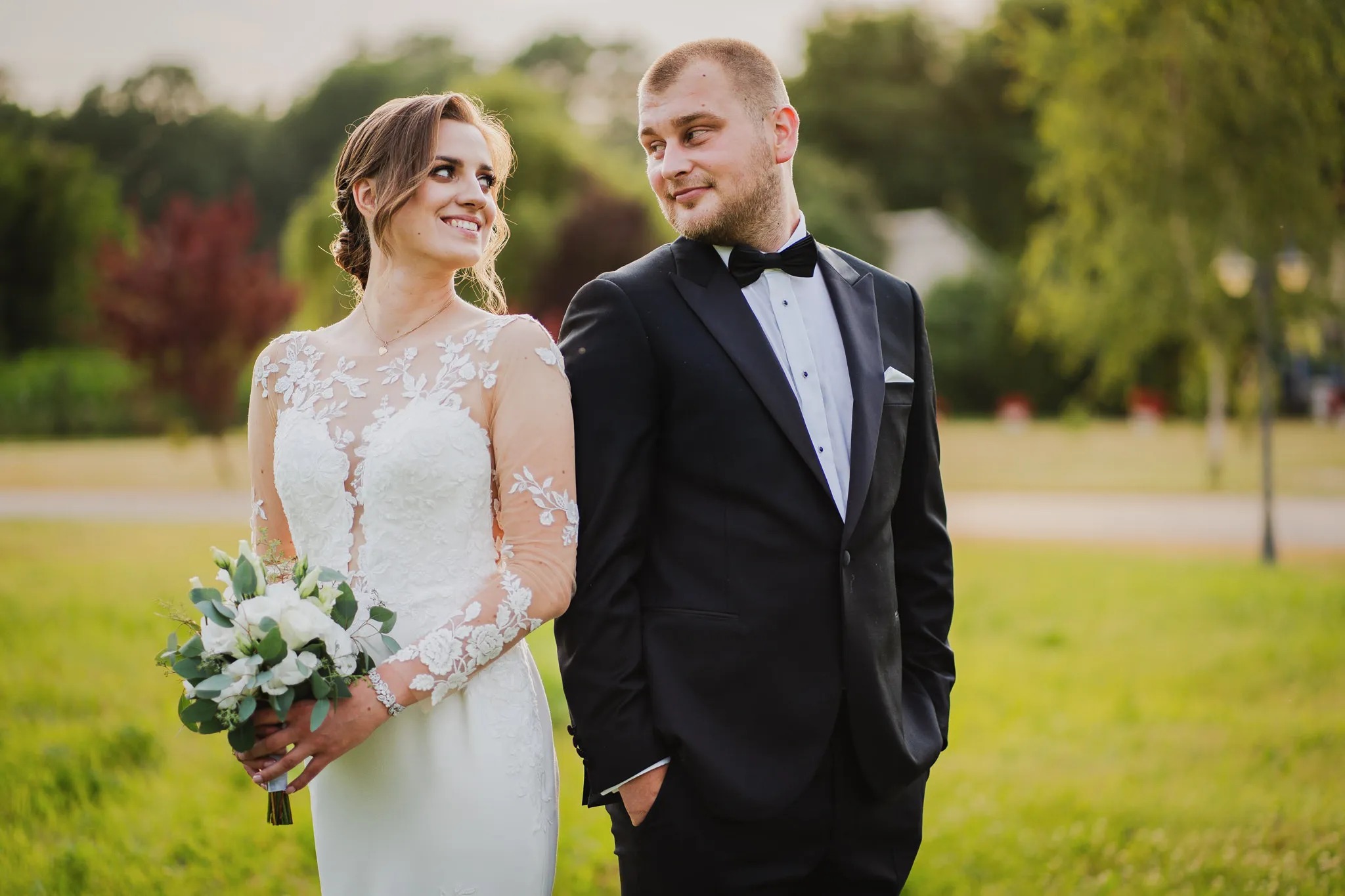 Uśmiechnięta panna młoda w koronkowej sukni trzyma bukiet, spoglądając na pana młodego w smokingu, uchwyconych podczas sesji w plenerze w Żyrardowie.