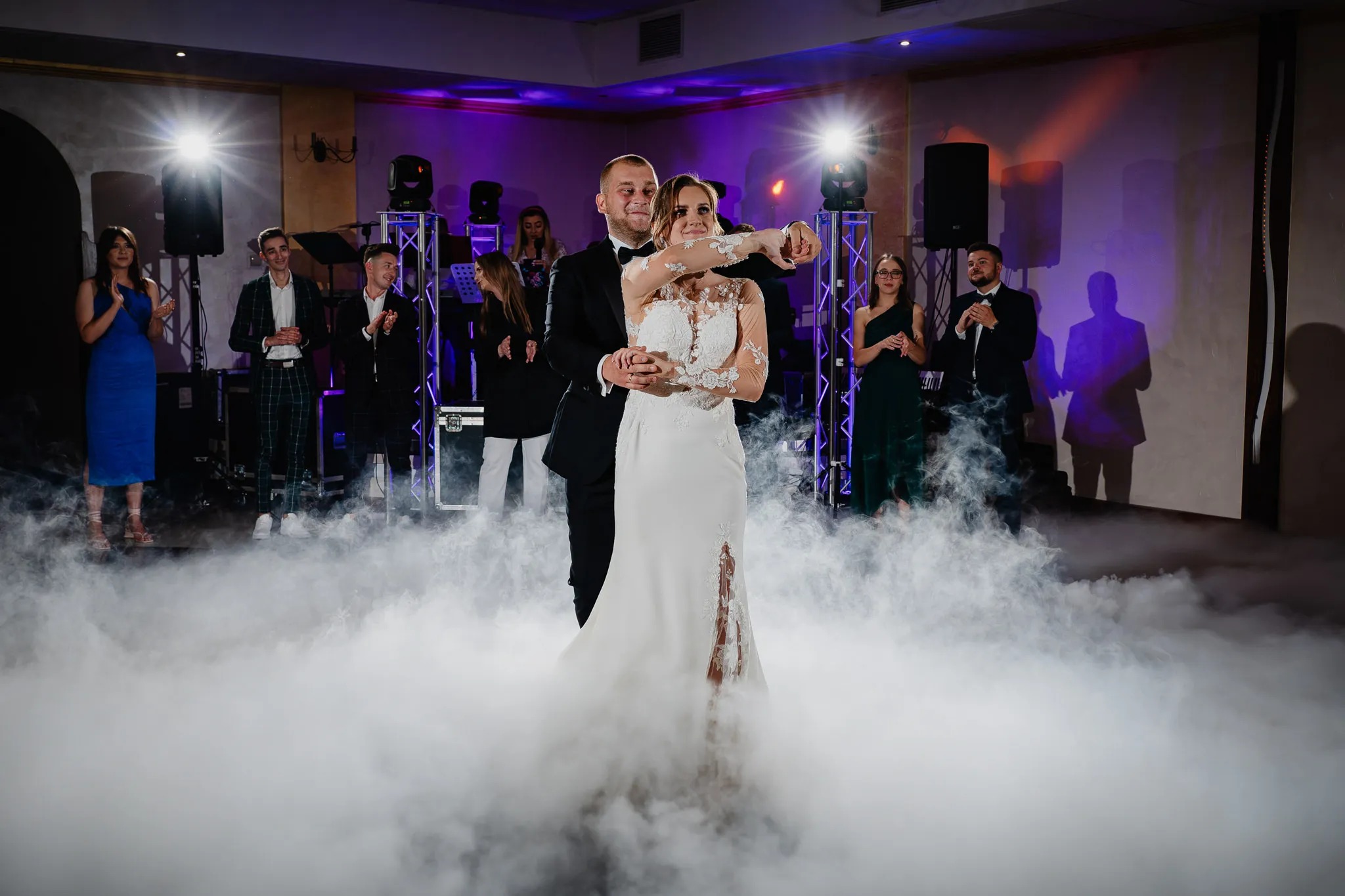 Para młoda tańczy pierwszy taniec w chmurze dymu, goście obserwują, oświetlenie sceniczne w tle.