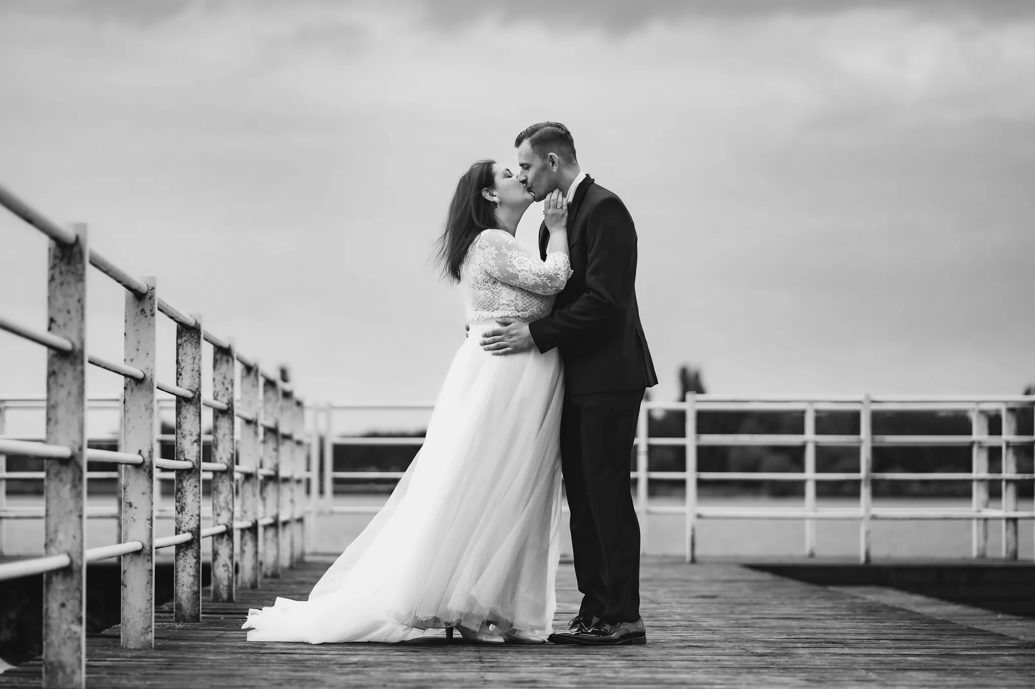 Czarno-białe zdjęcie całującej się pary młodej na drewnianym molo z metalowymi barierkami w tle, pochmurne niebo.