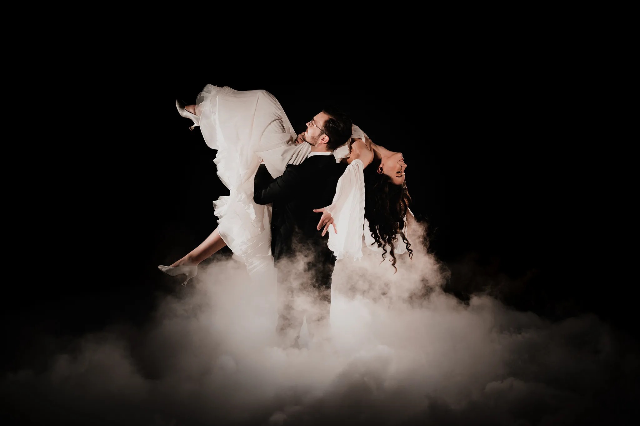 Pan młody w czarnym garniturze trzyma pannę młodą w białej sukni ślubnej, unosząc ją w powietrzu na tle dymu i czerni.