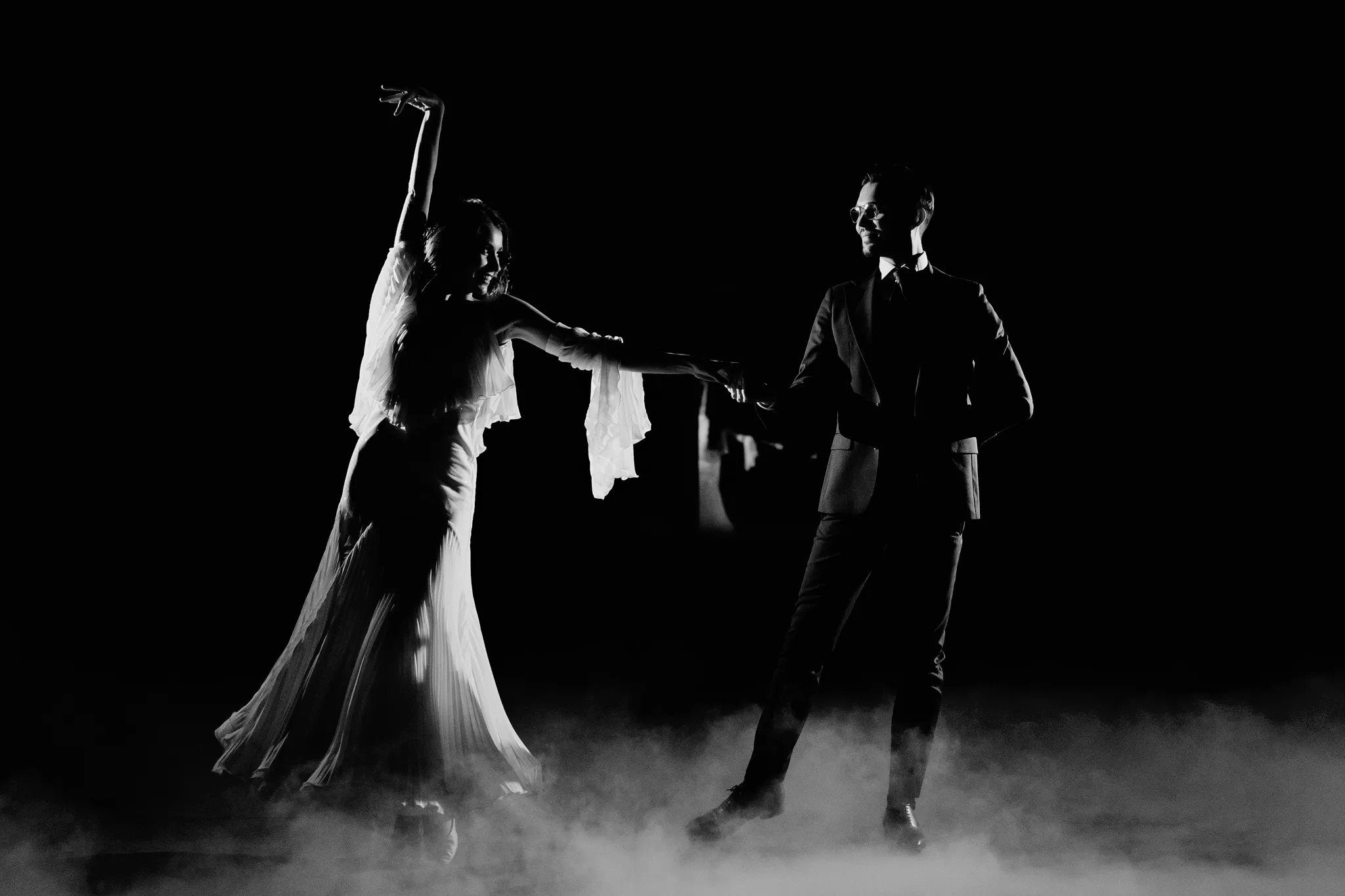 Para w strojach wieczorowych tańczy na scenie w dymie, czarno-białe ujęcie z niskim kluczem, ona w zwiewnej sukni z falbanami, on w garniturze i okularach.