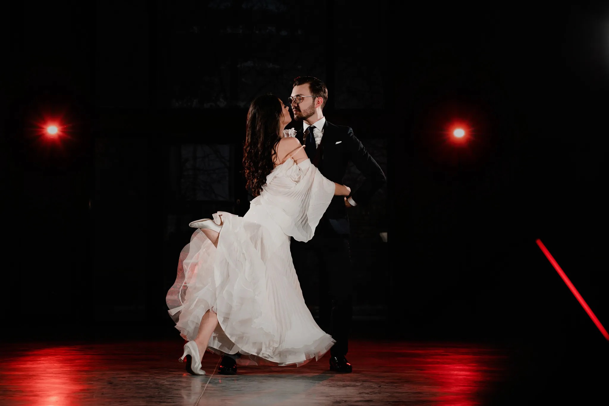 Para w tańcu: panna młoda w białej sukni z falbanami i pan młody w czarnym garniturze, podnoszona przez partnera na tle ciemnej sceny z czerwonymi reflektorami.