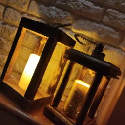 Dwa drewniane lampiony ze sznurkowymi uchwytami, jeden z białą, drugi ze złotą świecą, ustawione na marmurowym parapecie na tle ściany z dekoracyjnej cegły.