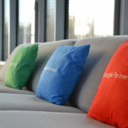 Trzy kolorowe poduszki (zielona, niebieska, czerwona) z napisem Google Partners leżą na szarej sofie na tle okna.