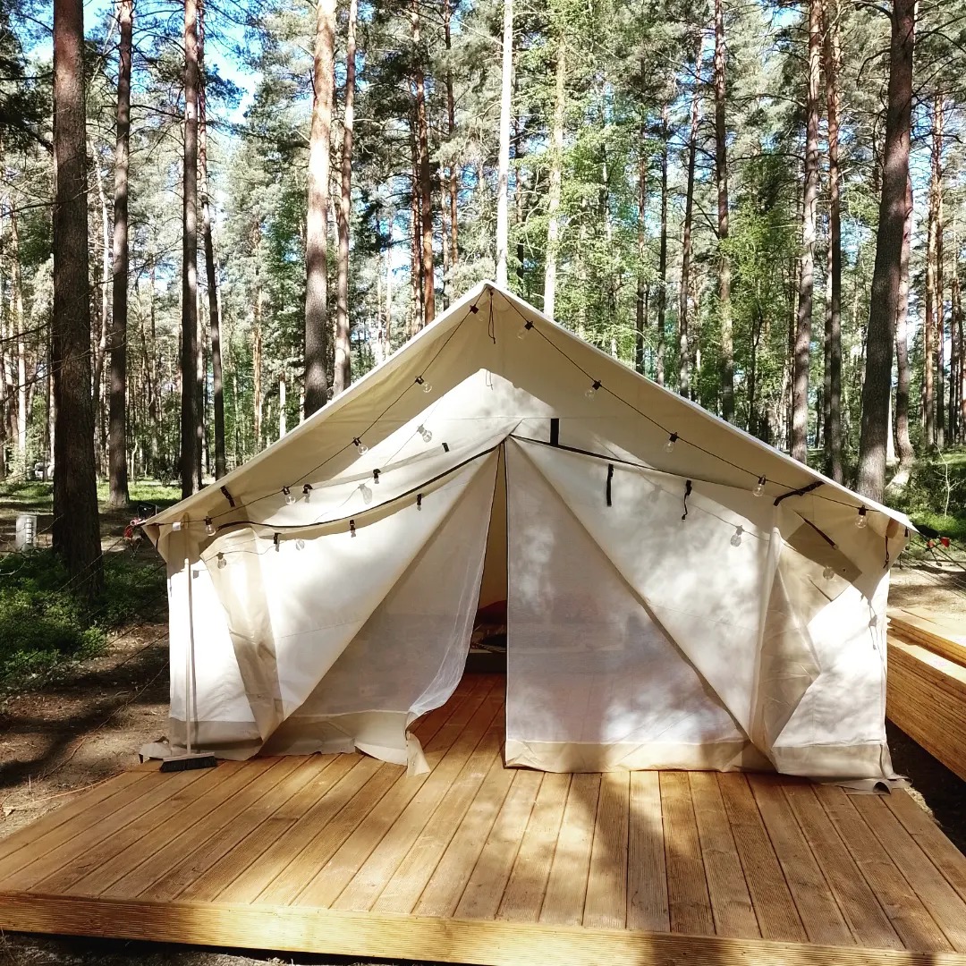 Biały namiot typu tipi z rozwieszonymi lampkami, ustawiony na drewnianym tarasie w lesie.