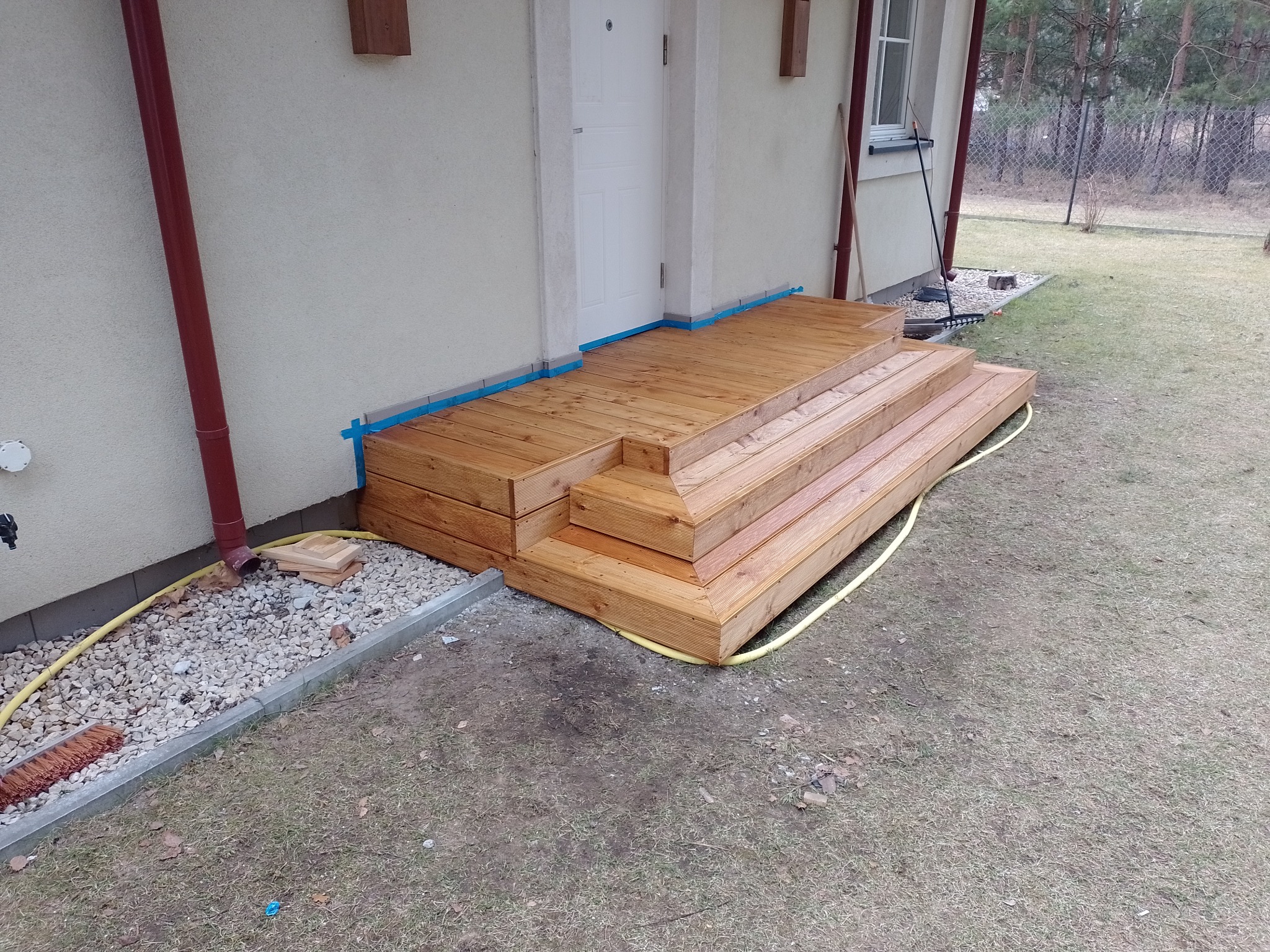 Nowo wybudowane drewniane schody wejściowe do domu, zabezpieczone niebieską taśmą malarską, z widocznym wężem ogrodowym i narzędziami w tle.
