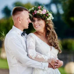 Para młoda w objęciach, panna młoda w białej sukni z koronkowymi rękawami i wiankiem z różowych i białych kwiatów, pan młody w białej koszuli z muszką, oboje patrzą na siebie z uśmiechem, tło zielone.