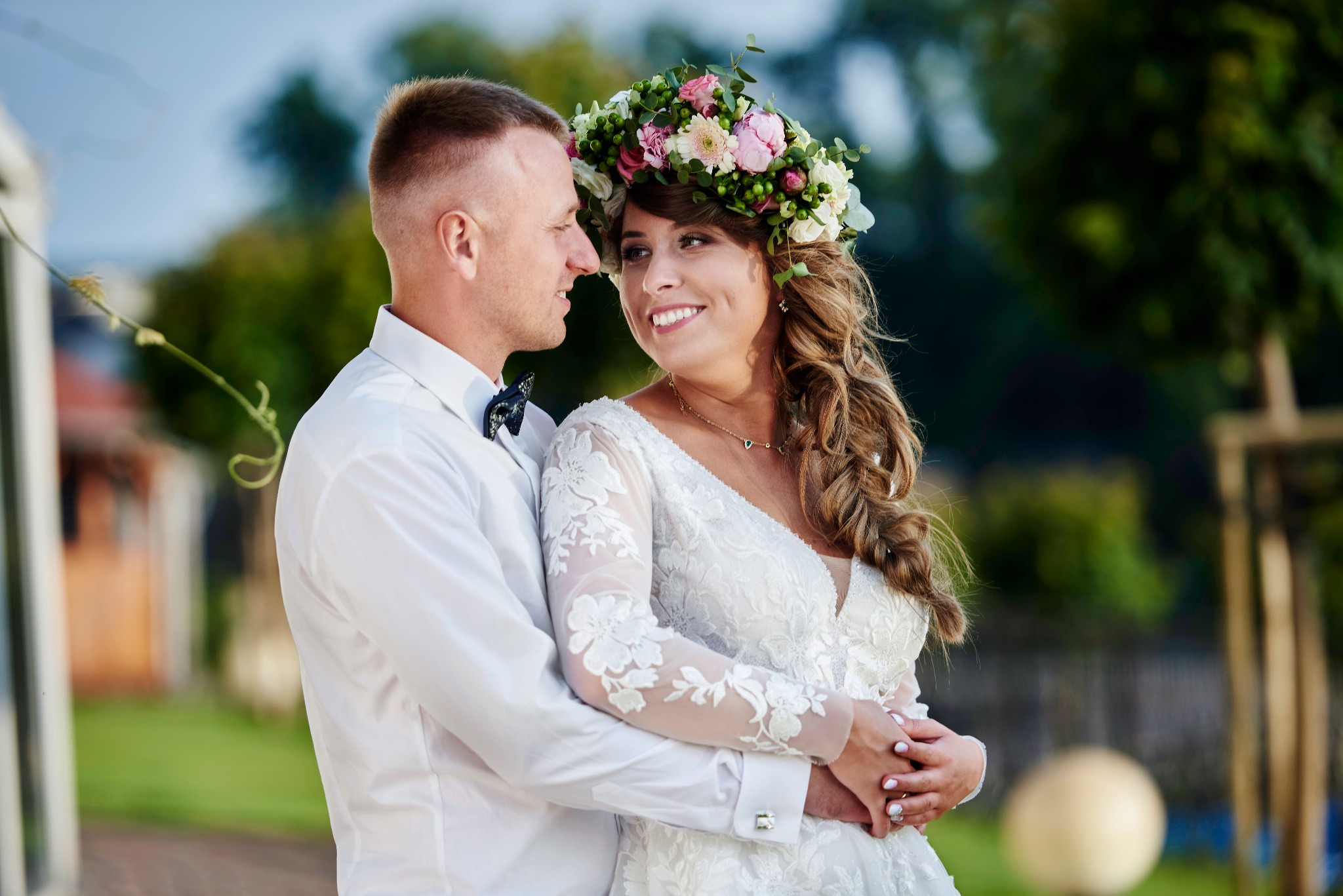 Para młoda w objęciach, panna młoda w białej sukni z koronkowymi rękawami i wiankiem z różowych i białych kwiatów, pan młody w białej koszuli z muszką, oboje patrzą na siebie z uśmiechem, tło zielone.