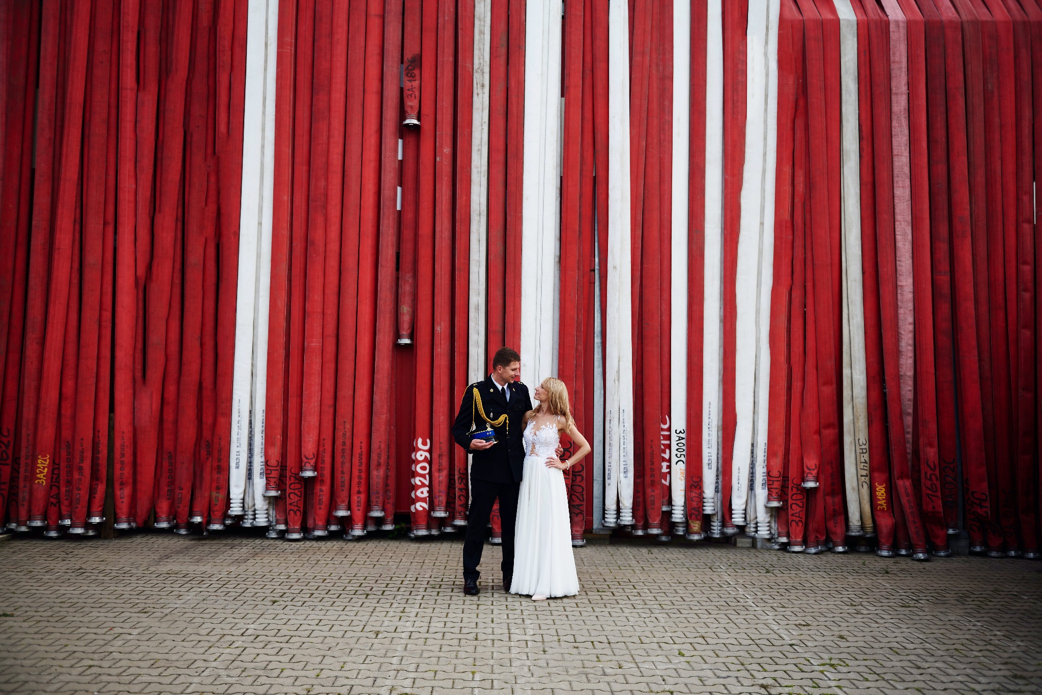 Para młoda: mężczyzna w galowym mundurze strażackim i kobieta w białej sukni ślubnej, pozują na tle ściany z czerwono-białych zwiniętych węży strażackich na brukowanym placu.