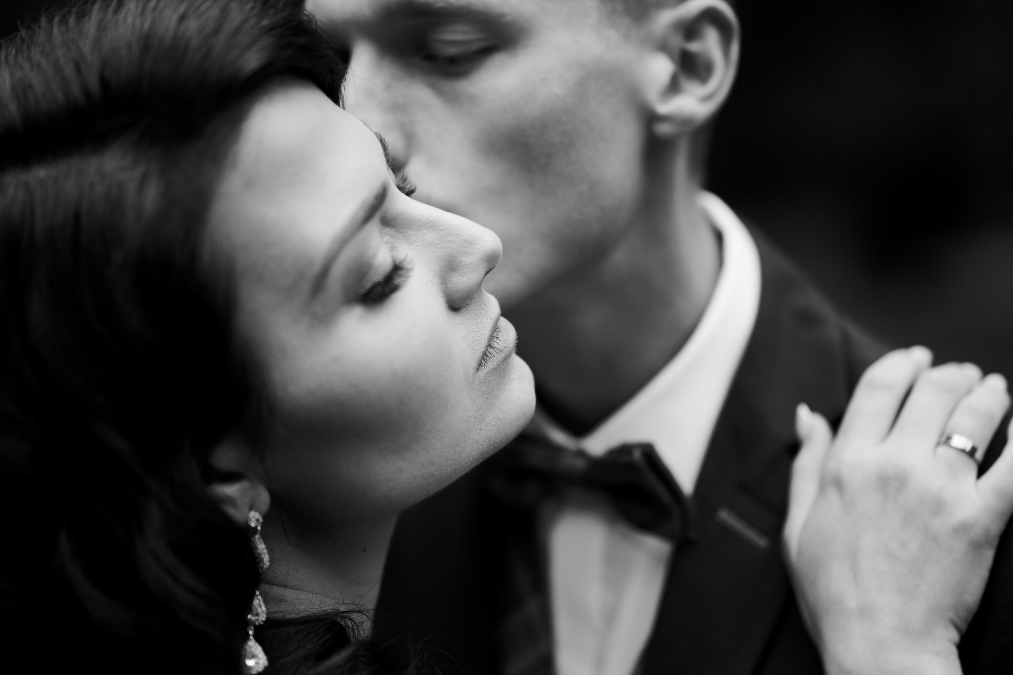 Czarno-białe zbliżenie: mężczyzna w garniturze całuje kobietę w szyję, widoczne długie kolczyki i kokarda.