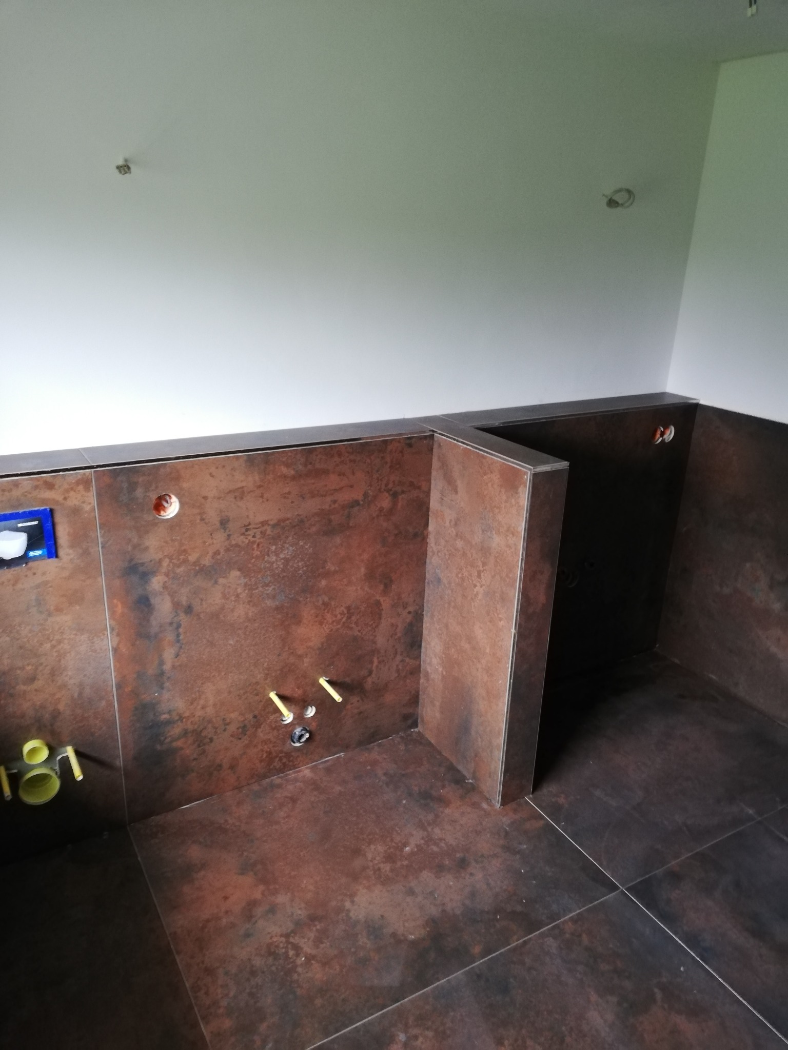 Wnętrze łazienki w trakcie wykańczania, z brązowymi płytkami imitującymi rdzę na ścianach i podłodze. Widoczne wystające rury instalacji wodnej i kanalizacyjnej oraz otwory na oświetlenie w suficie.
