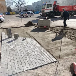 Fragment placu z częściowo ułożoną szarą kostką brukową, paleta z kostką, ubijarka i pracownik wyrównujący podłoże, w tle zaparkowane samochody i budynek mieszkalny.