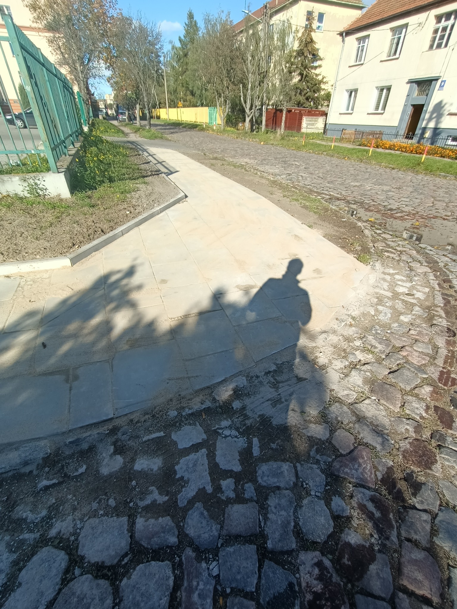 Nowo ułożony chodnik z jasnych płyt, kontrastujący z fragmentem starej, nierównej drogi z kamiennej kostki. Widoczny cień osoby robiącej zdjęcie i fragment zielonego ogrodzenia w tle.