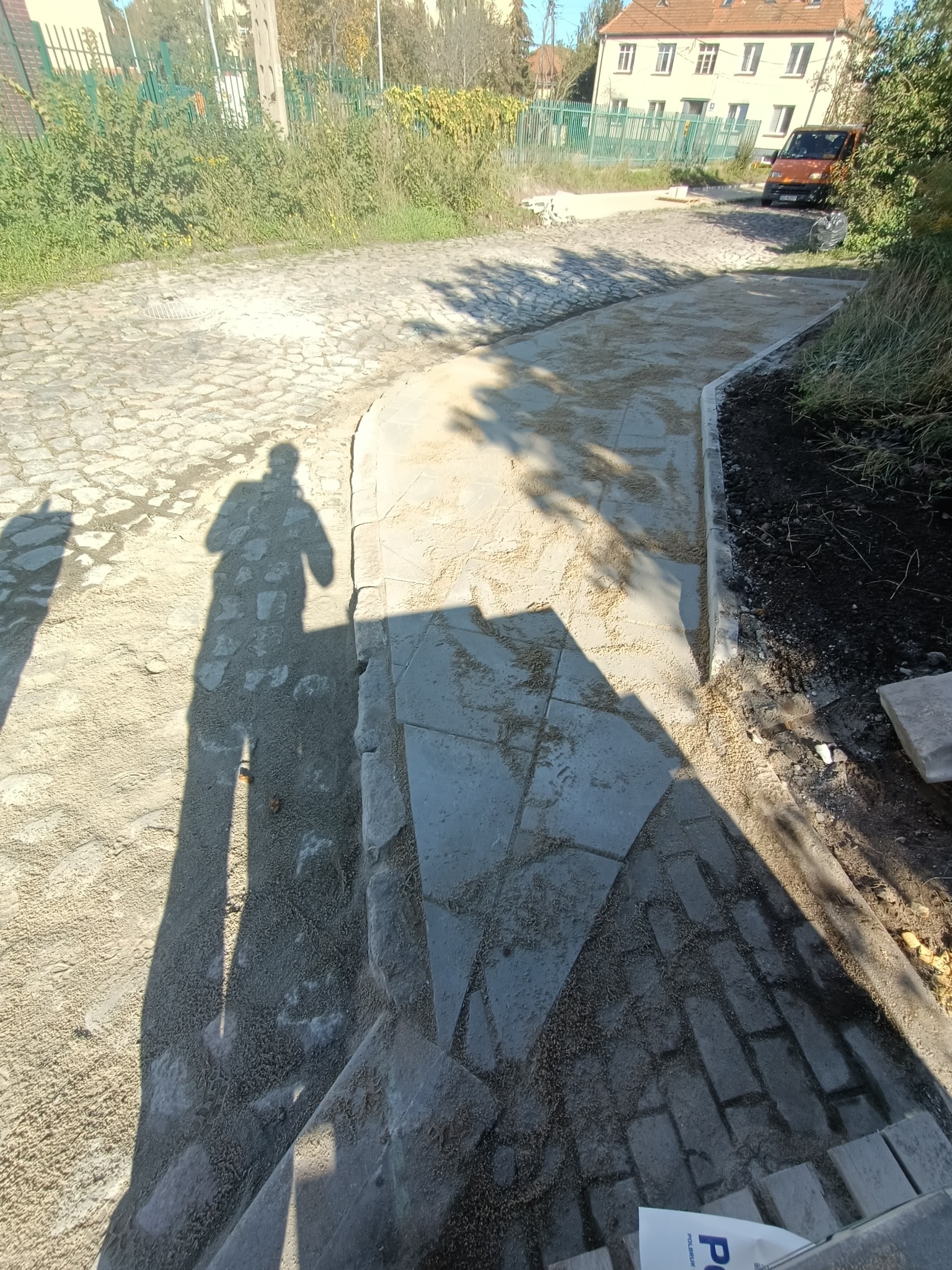 Fragment drogi z kostki brukowej w trakcie budowy, widoczny cień osoby, podsypka piaskowa i obrzeża, w tle zabudowania i zieleń.
