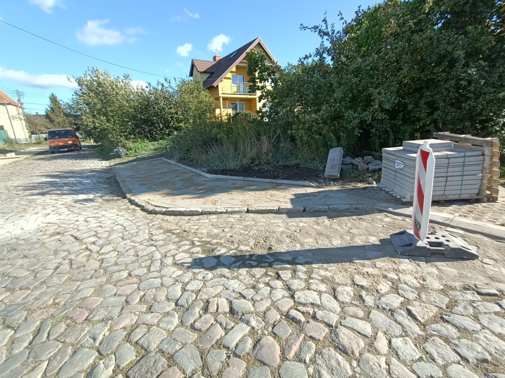 Fragment placu z brukowaną nawierzchnią, częściowo wyłożoną nową kostką; w tle widoczny dom z żółtą elewacją, drzewa oraz pomarańczowy samochód dostawczy.