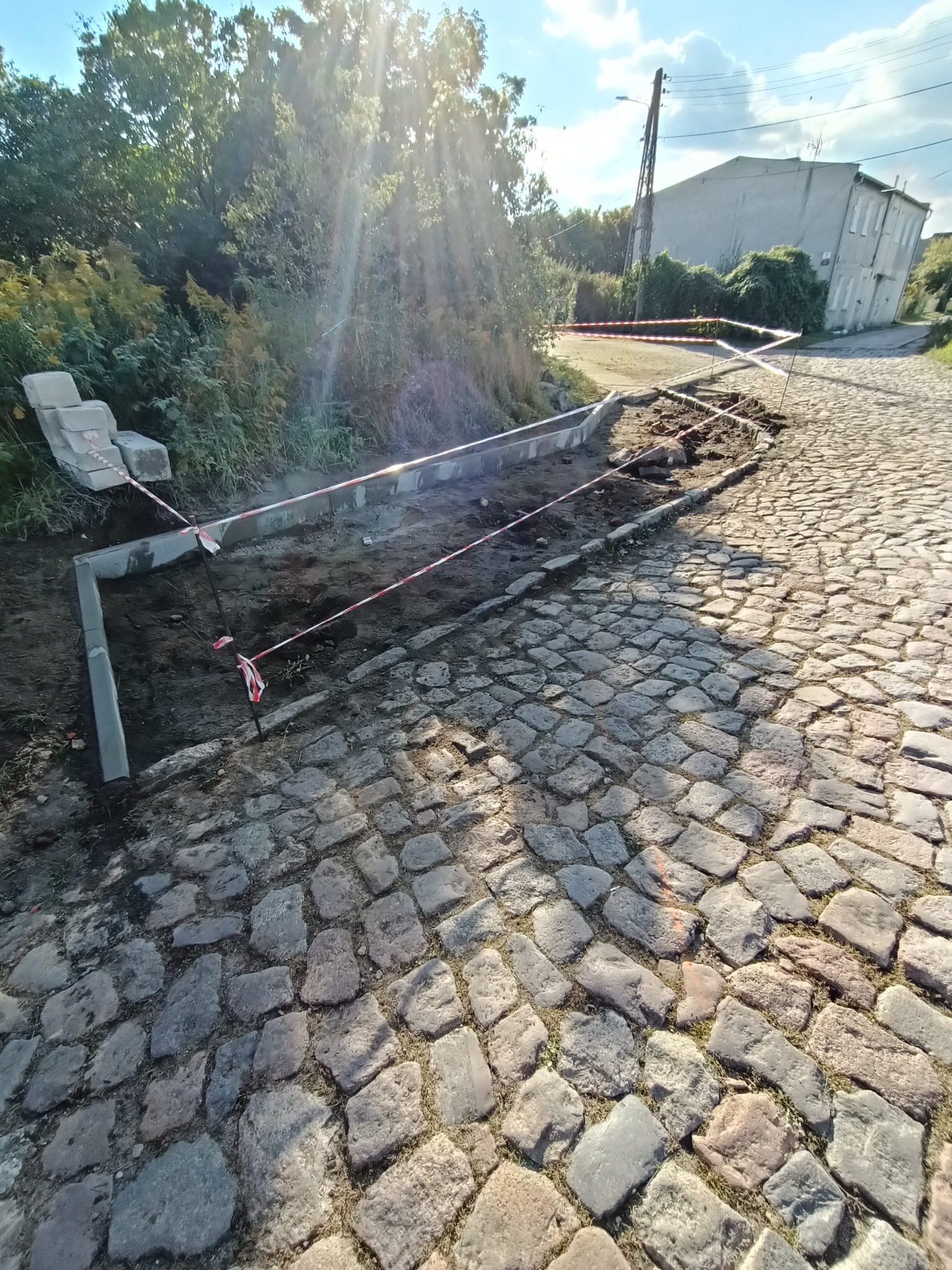 Układanie obrzeży betonowych na drodze z kostki brukowej, zabezpieczone taśmą ostrzegawczą, w tle budynek i drzewa w słoneczny dzień.