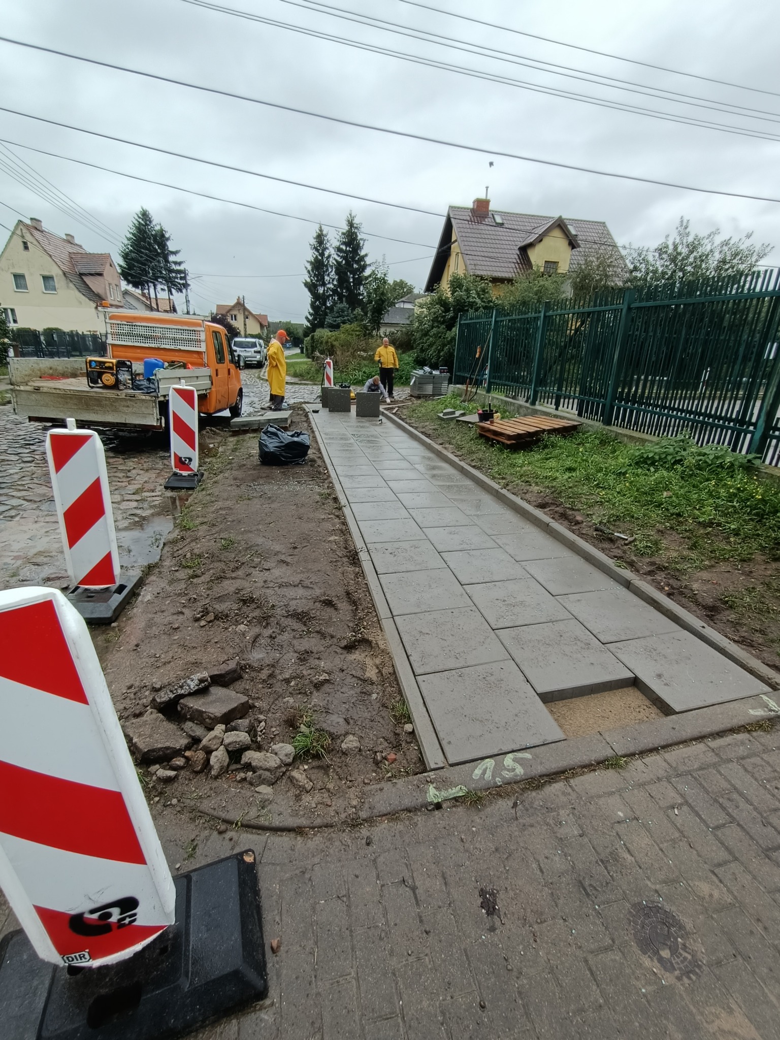Montaż chodnika z betonowych płyt na przygotowanym podłożu piaskowym, widoczne narzędzia, ekipa w odblaskowych ubraniach i pomarańczowy samochód dostawczy, zabezpieczenia budowy w postaci słupków...