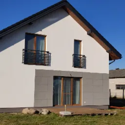 Nowoczesny dom jednorodzinny z białą elewacją, szarym cokołem z płytek elewacyjnych, drewnianą stolarką okienną i czarnymi balustradami balkonowymi, widoczny w słoneczny dzień na tle błękitnego...